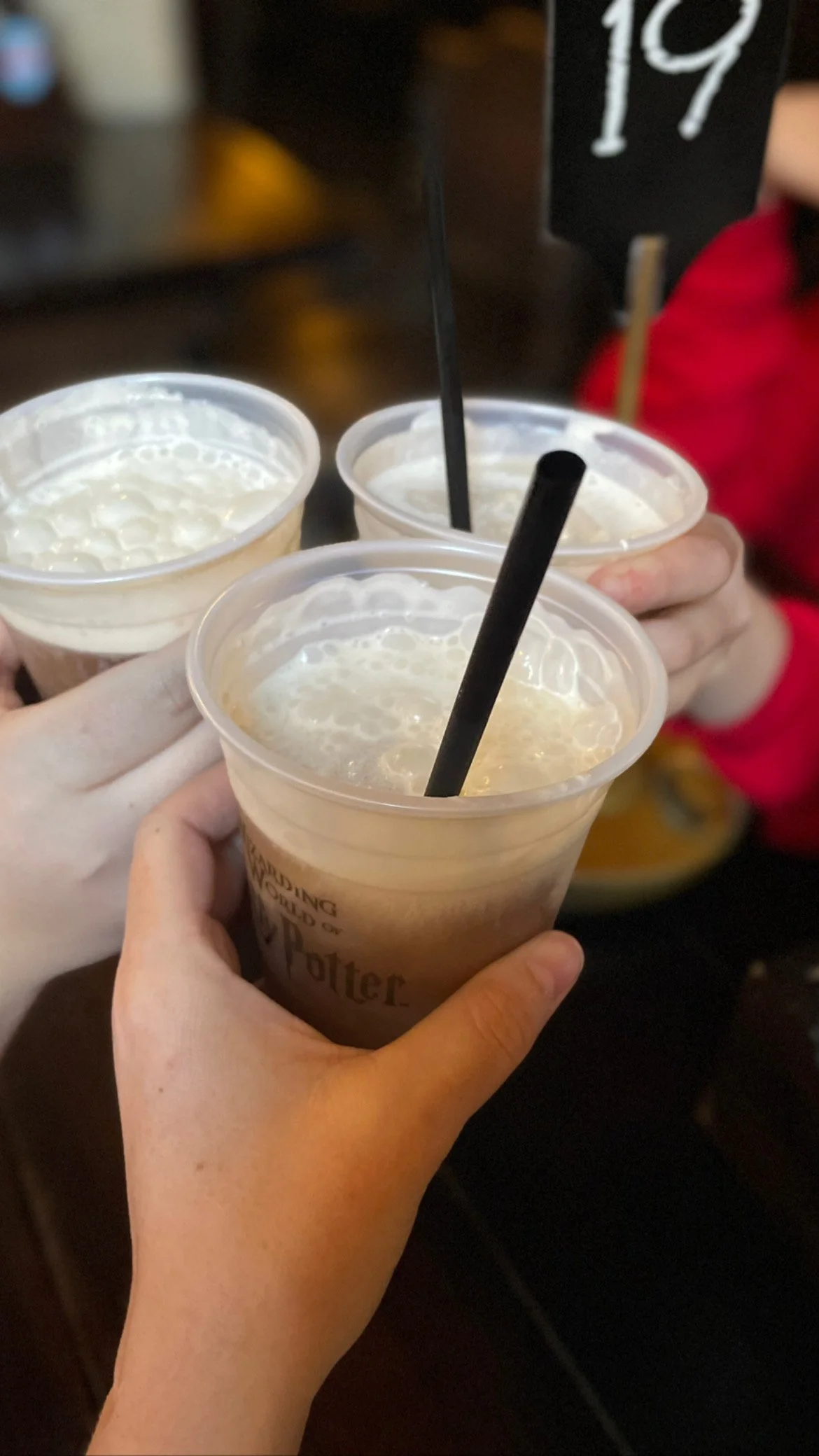 Three people holding up glasses of butterbeer from Harry Potter, with whipped cream topping, in a dark setting.