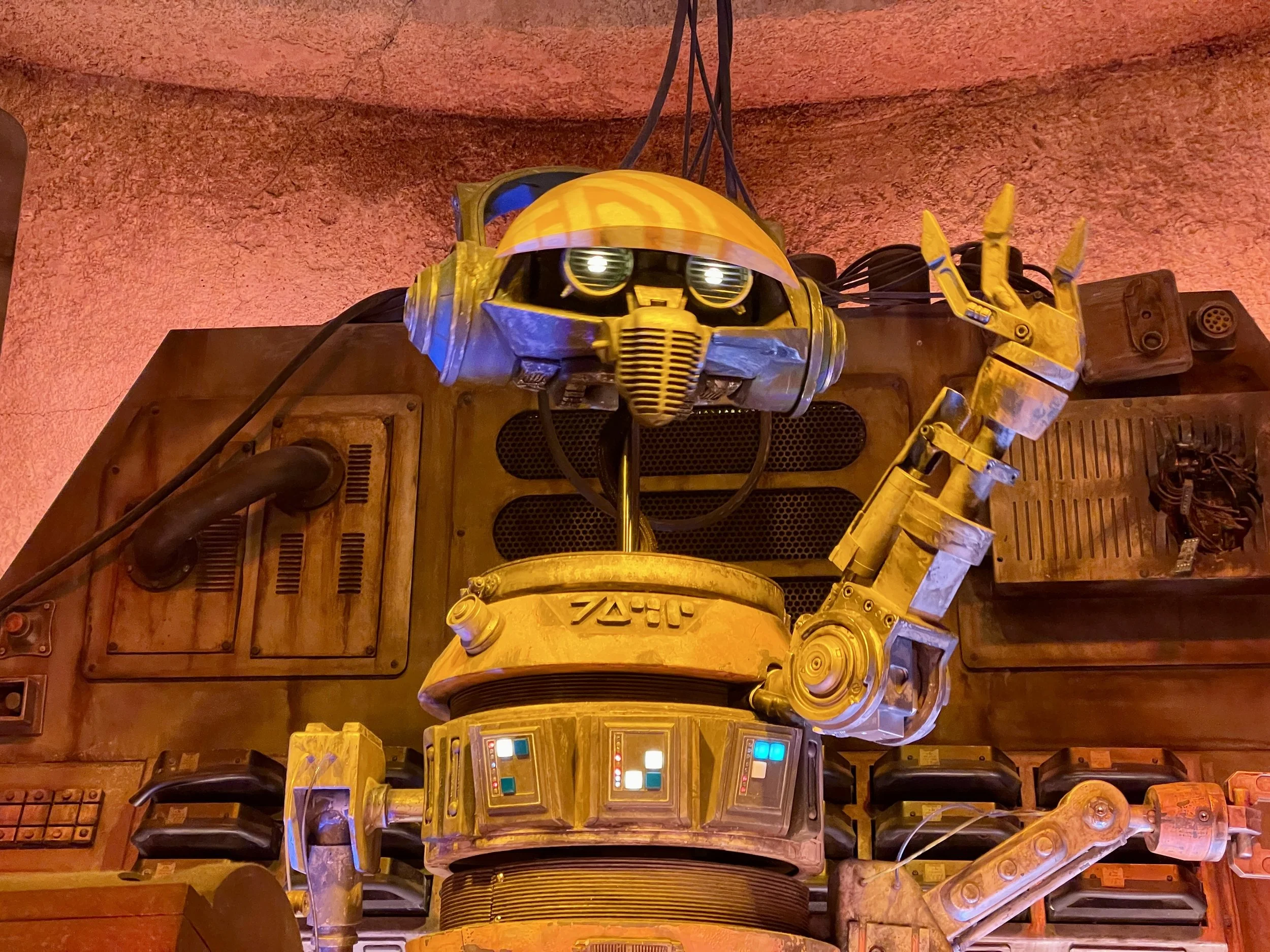 Close-up of a robot resembling WALL-E from the animated film, with a yellow and black color scheme, set against a background of wooden and metal objects, with a curved yellow head and big expressive eyes.