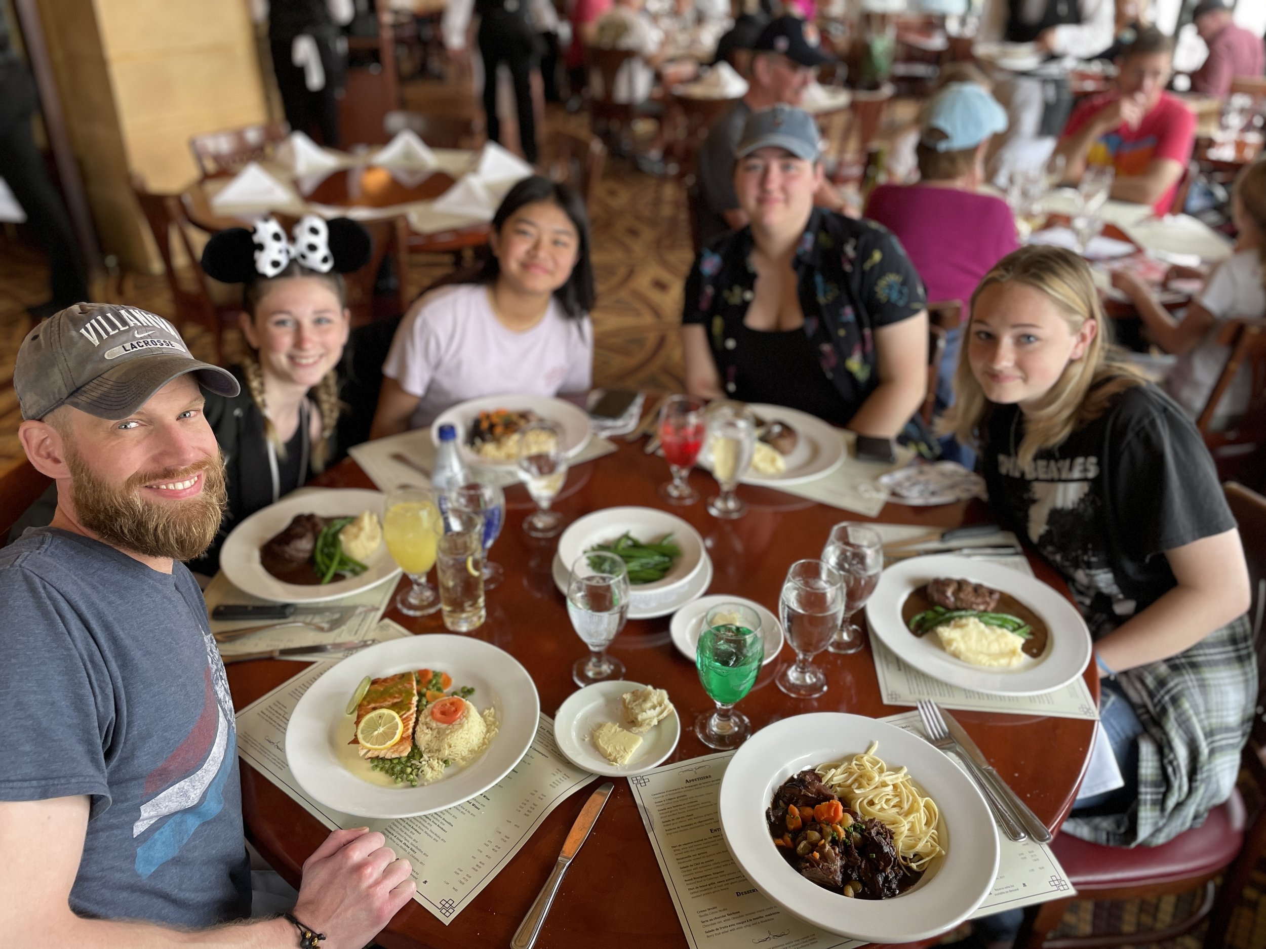 A group of six people, including a man with a beard and five young women, sitting around a round dining table in a restaurant, with food and drinks on the table.