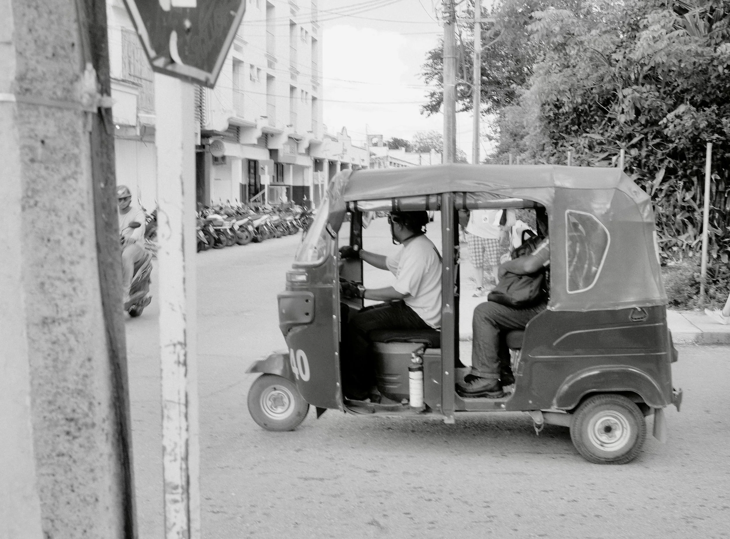 Black and white photo of a tuk-tuk vehicle on a city street, with two men inside, one driving and one seated with arms crossed, and a person in the background riding a scooter.