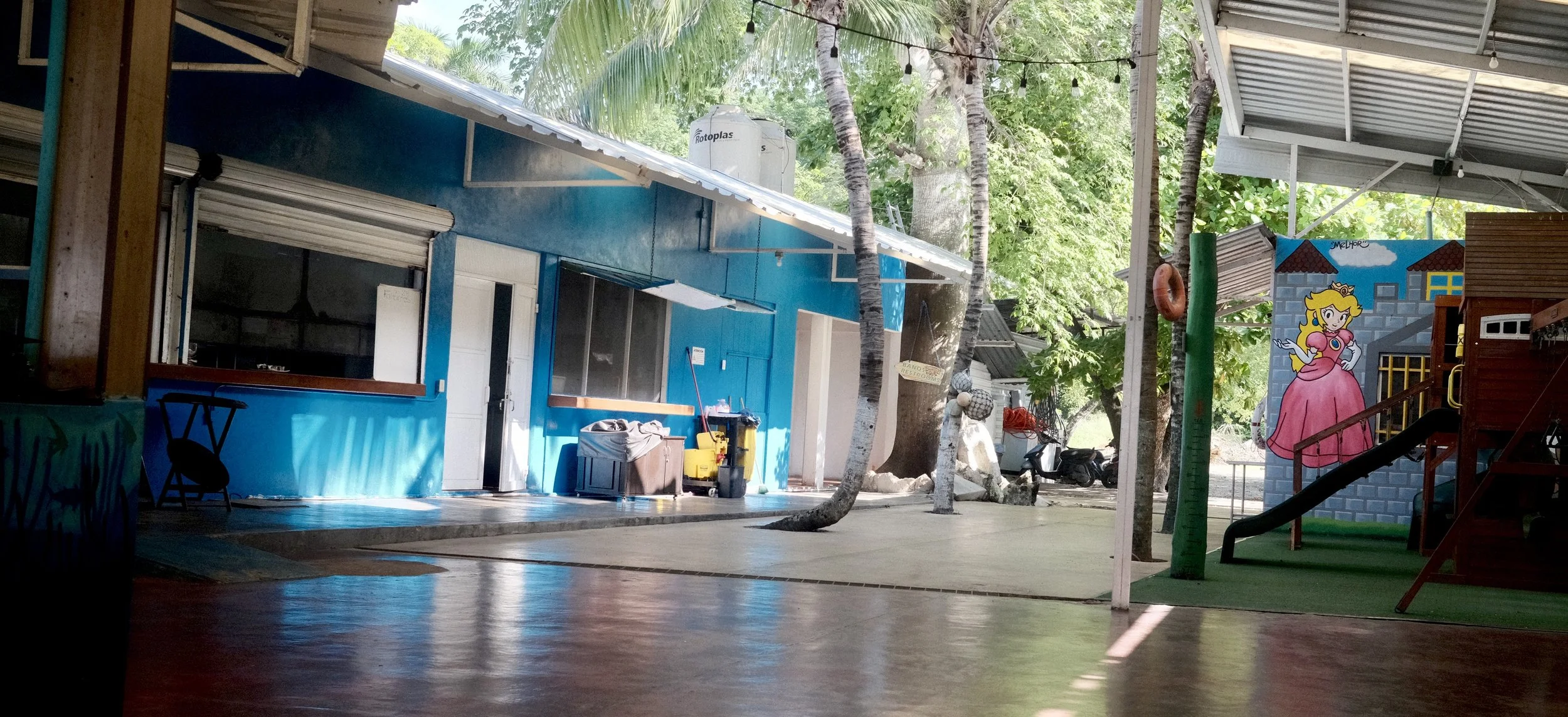 An open-air space with a blue building, trees, and cartoon princess mural, featuring a slide, inflatable structure, and playground equipment.