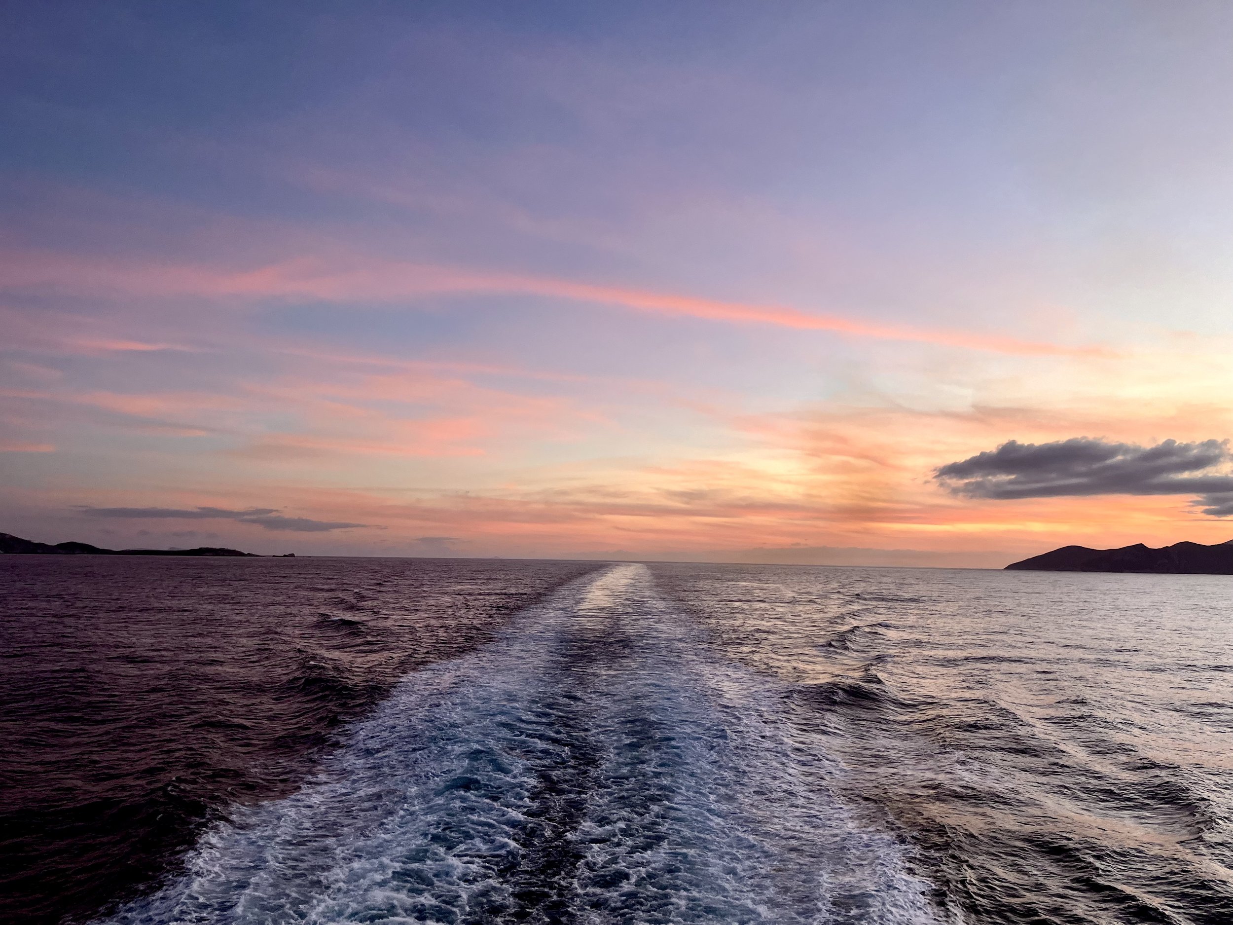 A view of the ocean during sunset with a boat trail in the water and pink, orange, and purple hues in the sky.