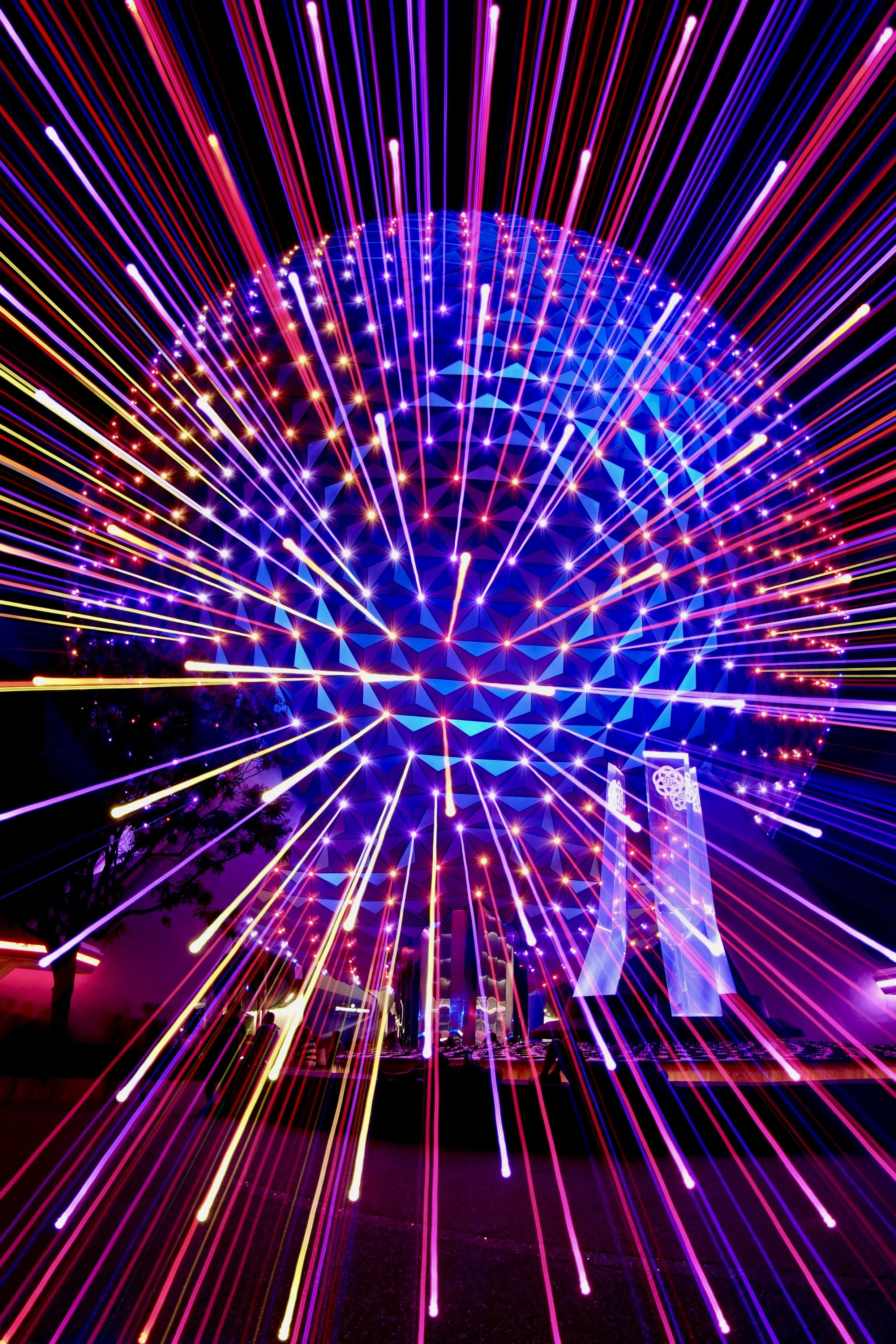 Colorful fireworks display at night with illuminated structures in the background, including two tall, translucent towers.