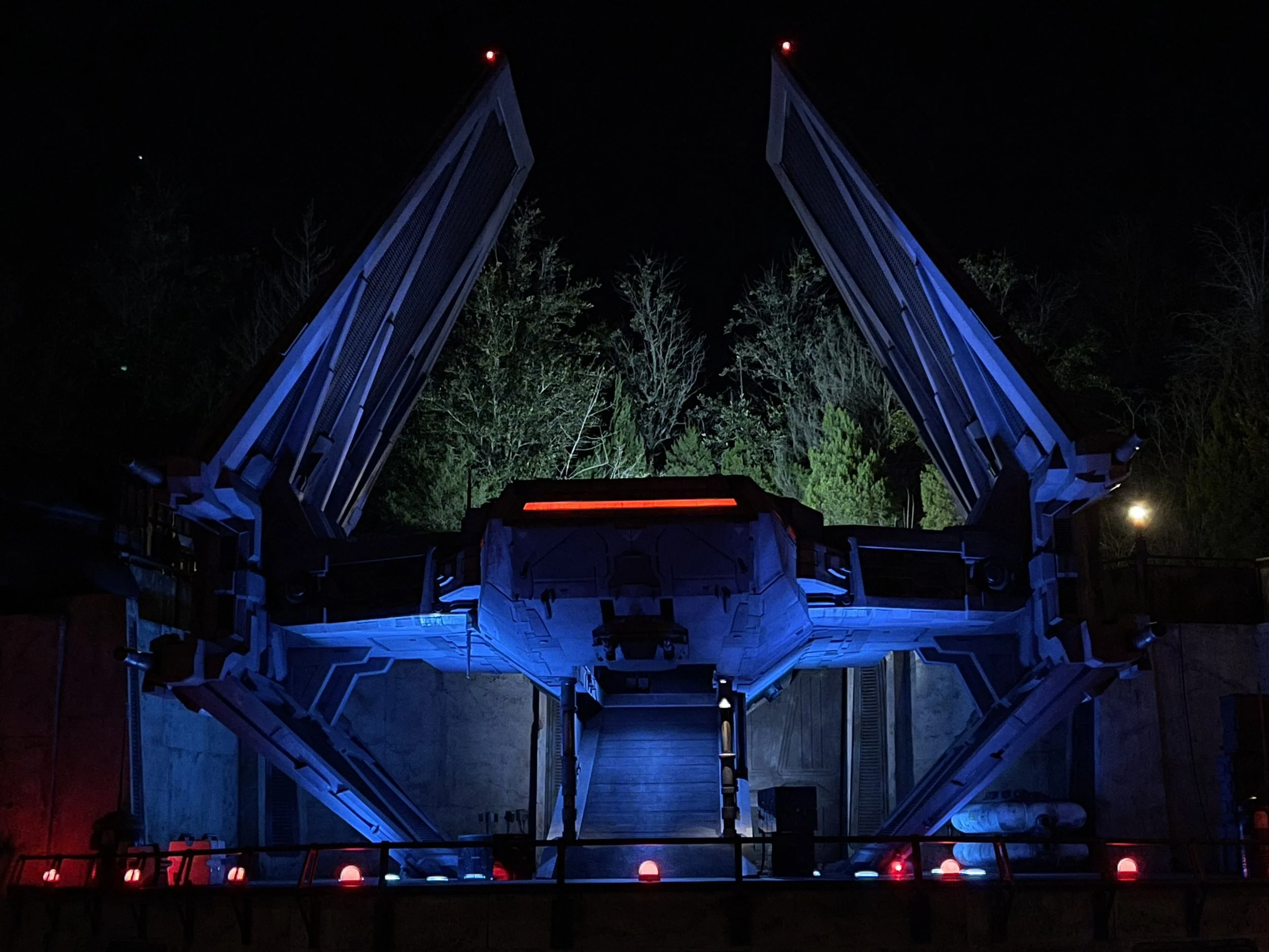 A Star Wars-themed spaceship launch pad at night with overcast sky and trees in the background, illuminated by blue and red lights.