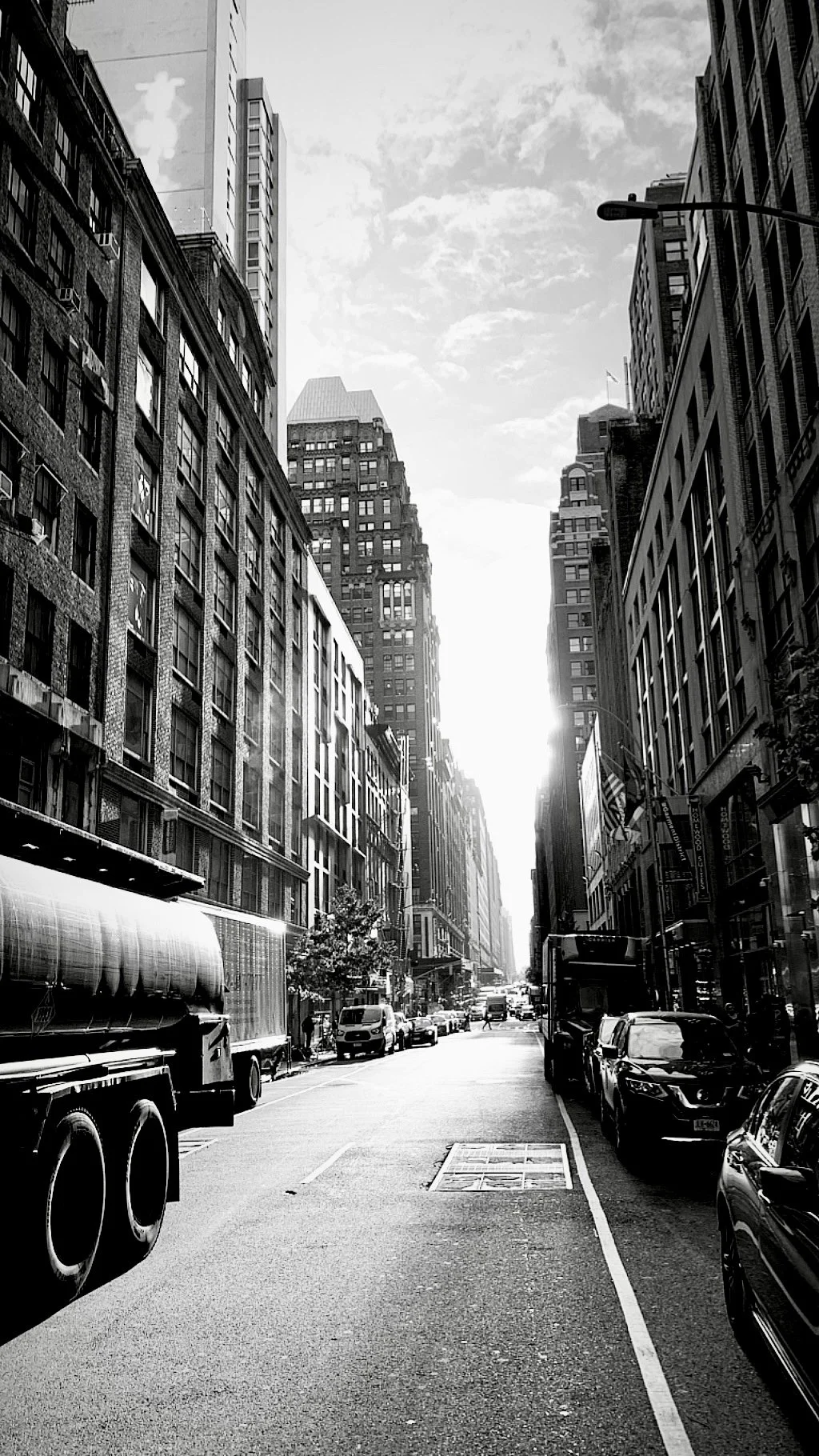 Black and white photo of a city street with tall buildings lining both sides, cars parked along the curb, and the sun setting or rising between the buildings.