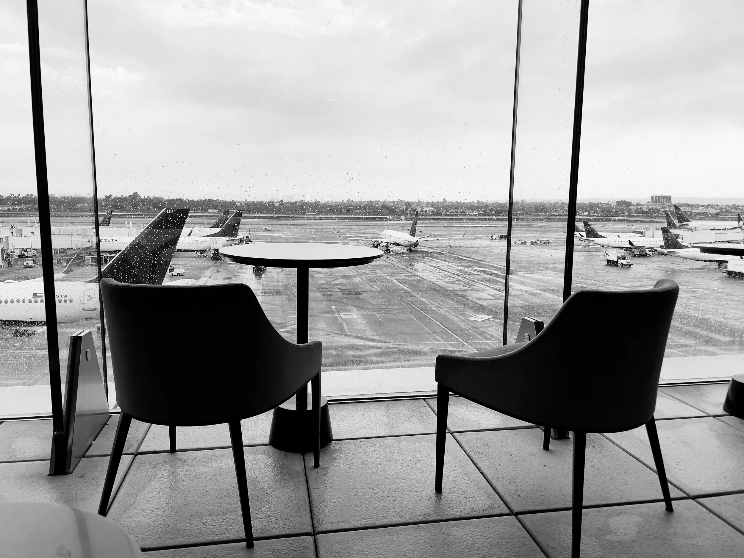 Airport terminal with two chairs and a small round table, view of airplanes parked on the tarmac through the rain-dappled glass window.
