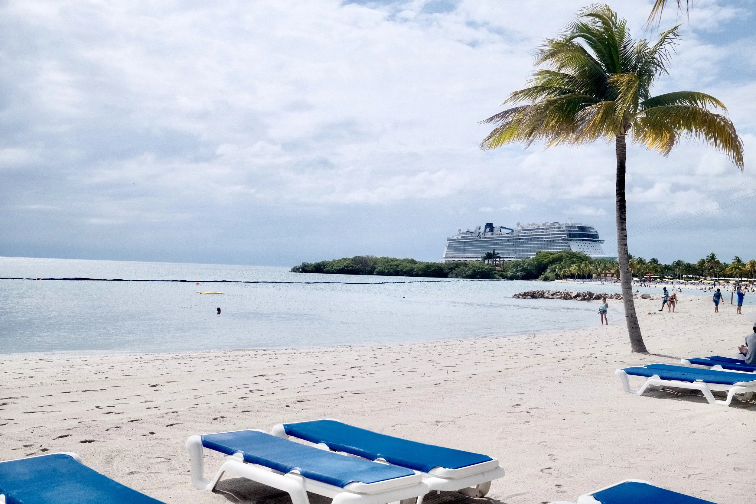 Beach with lounge chairs, palm trees, people, and a cruise ship in the distance on a cloudy day.