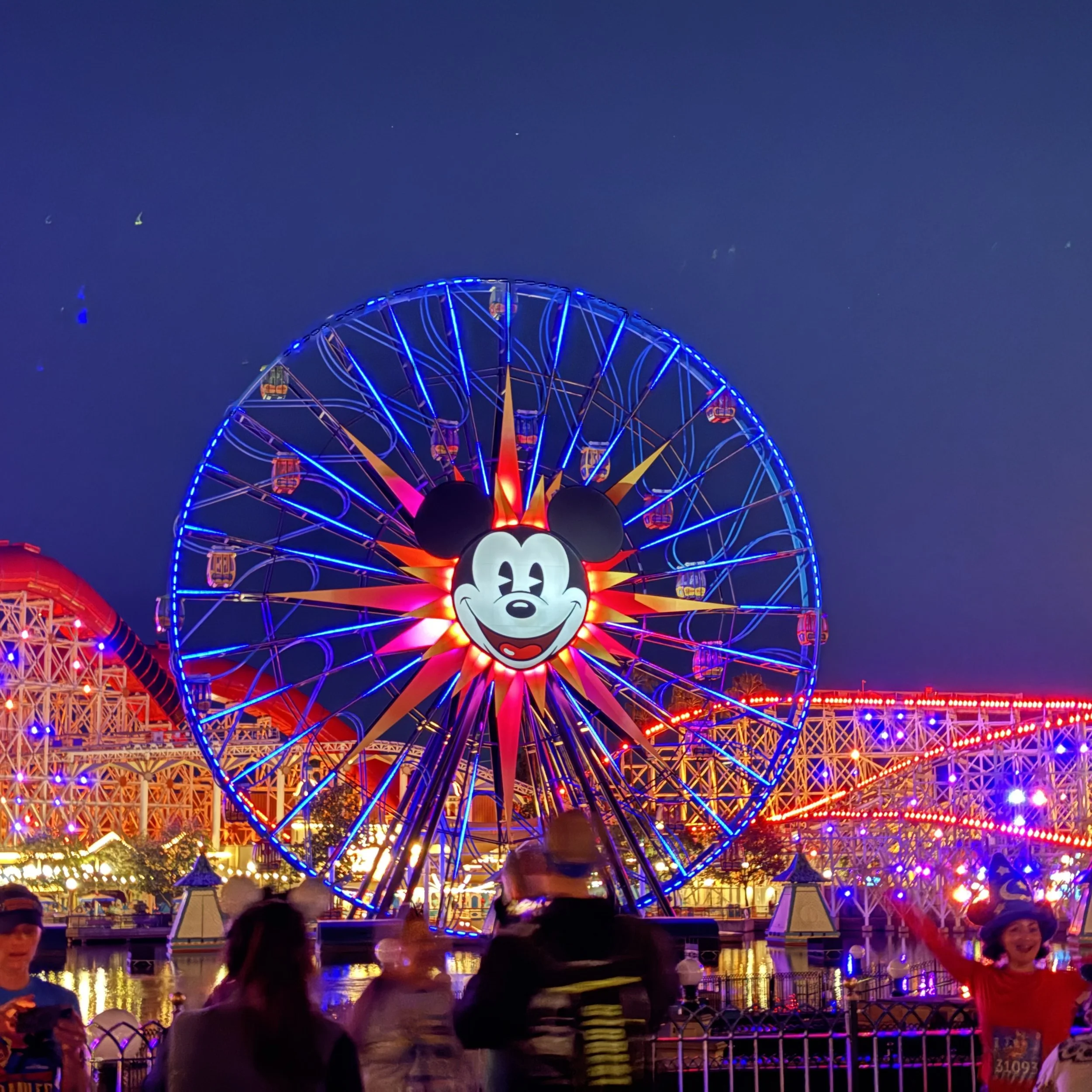 Nighttime scene at a Disney theme park with a large illuminated Mickey Mouse Ferris wheel in the background. People are walking and taking photos in front of the wheel, with colorful lights and a roller coaster visible in the distance.
