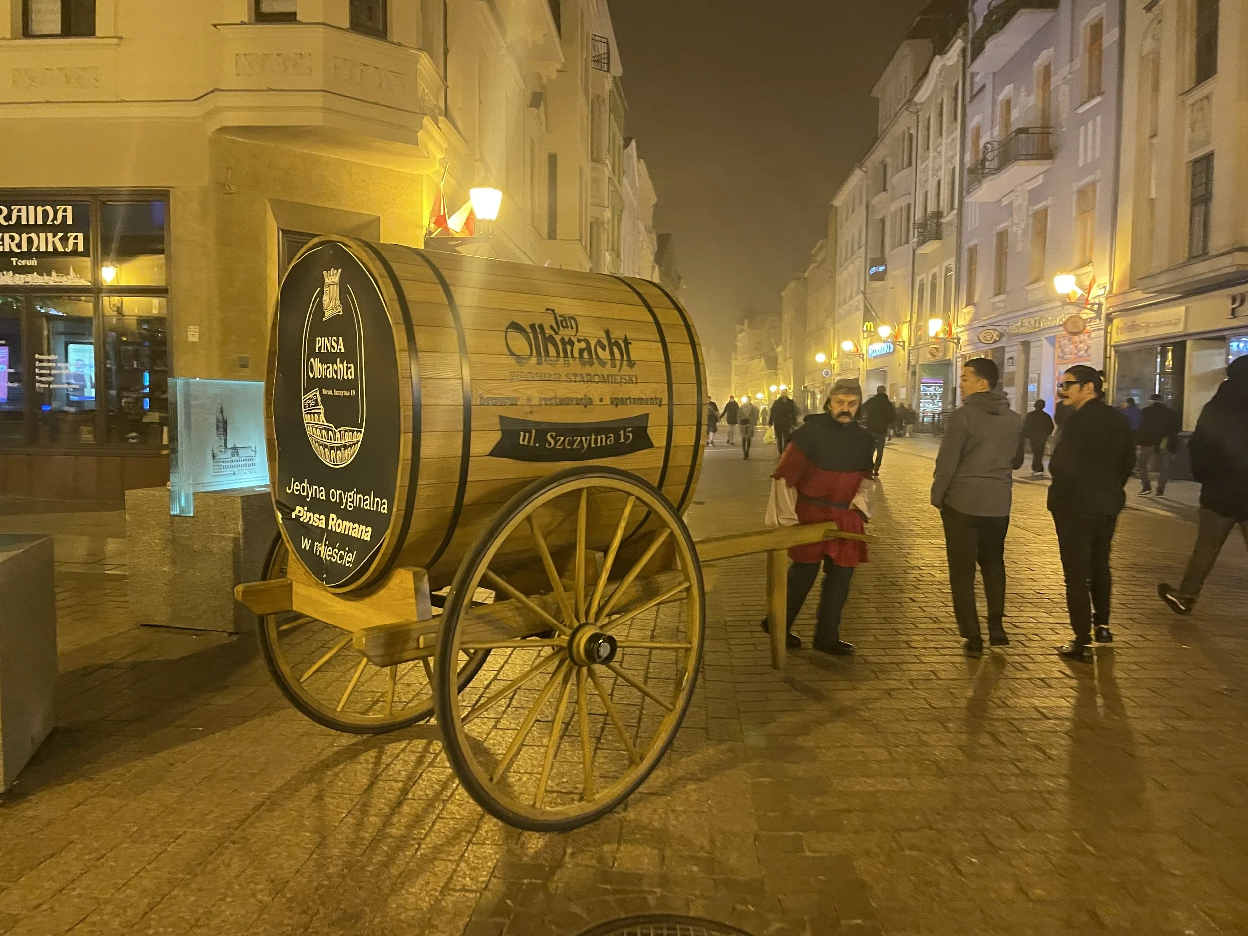 Night scene of a cobblestone street with people walking, illuminated by warm yellow streetlights. A wooden cart with a black sign, advertising Pineza Olbricht and mentioning its address and Roman origin, is in the foreground. The street is lined with