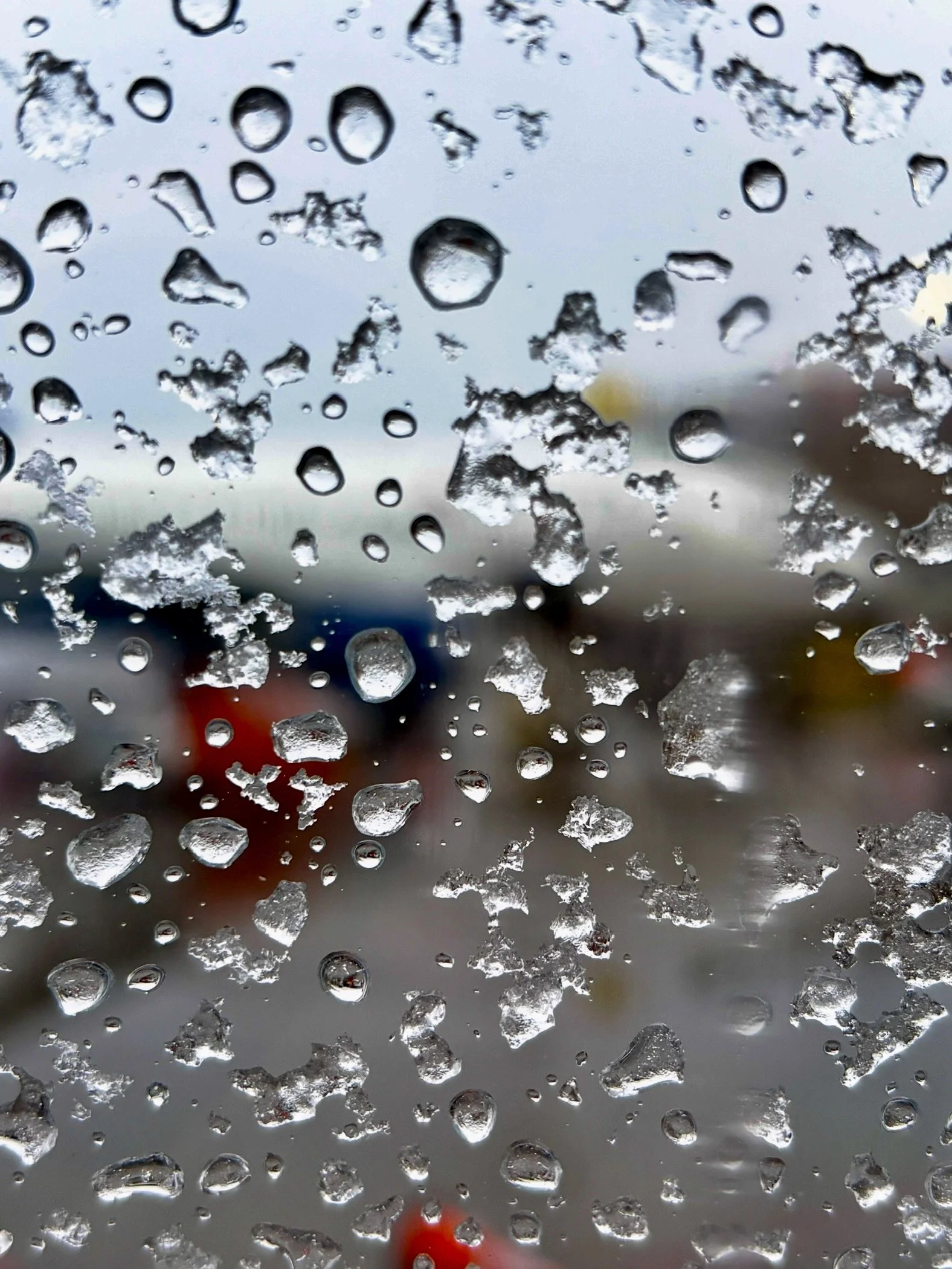 Rain droplets on a window with a blurred background of a cloudy sky and vehicles.