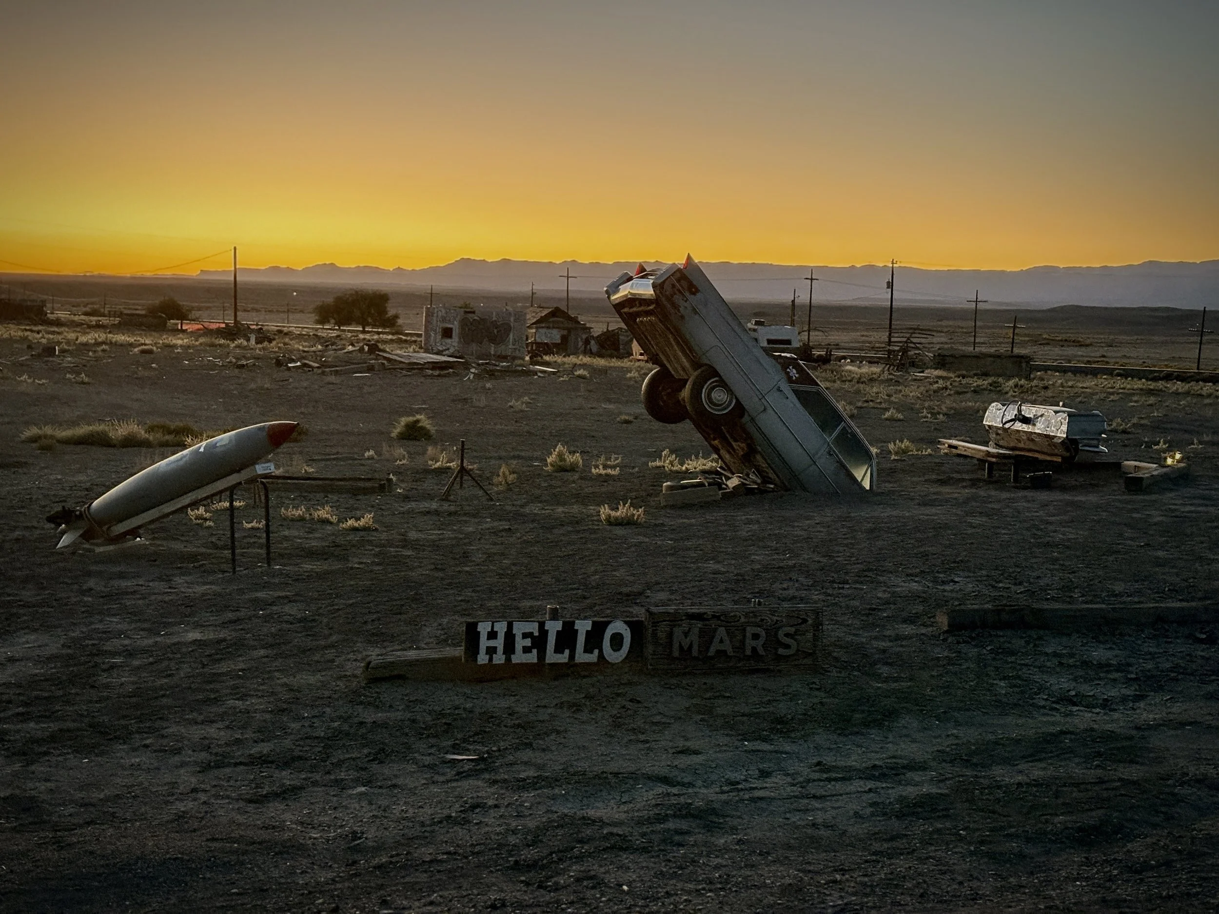 Desert with a sunset, a crashed vintage car, a rocket, and a sign that reads 'HELLO MARS'