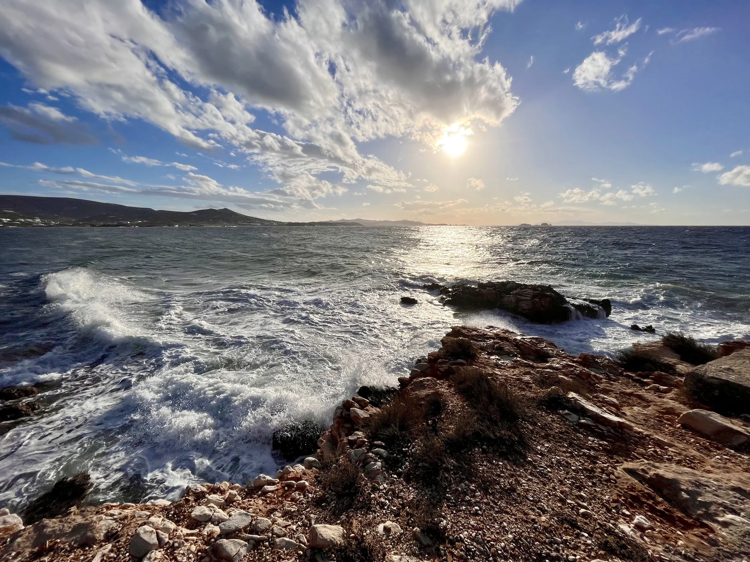 View of the ocean with waves crashing against rocks on the shore, under a partly cloudy sky with the sun shining.