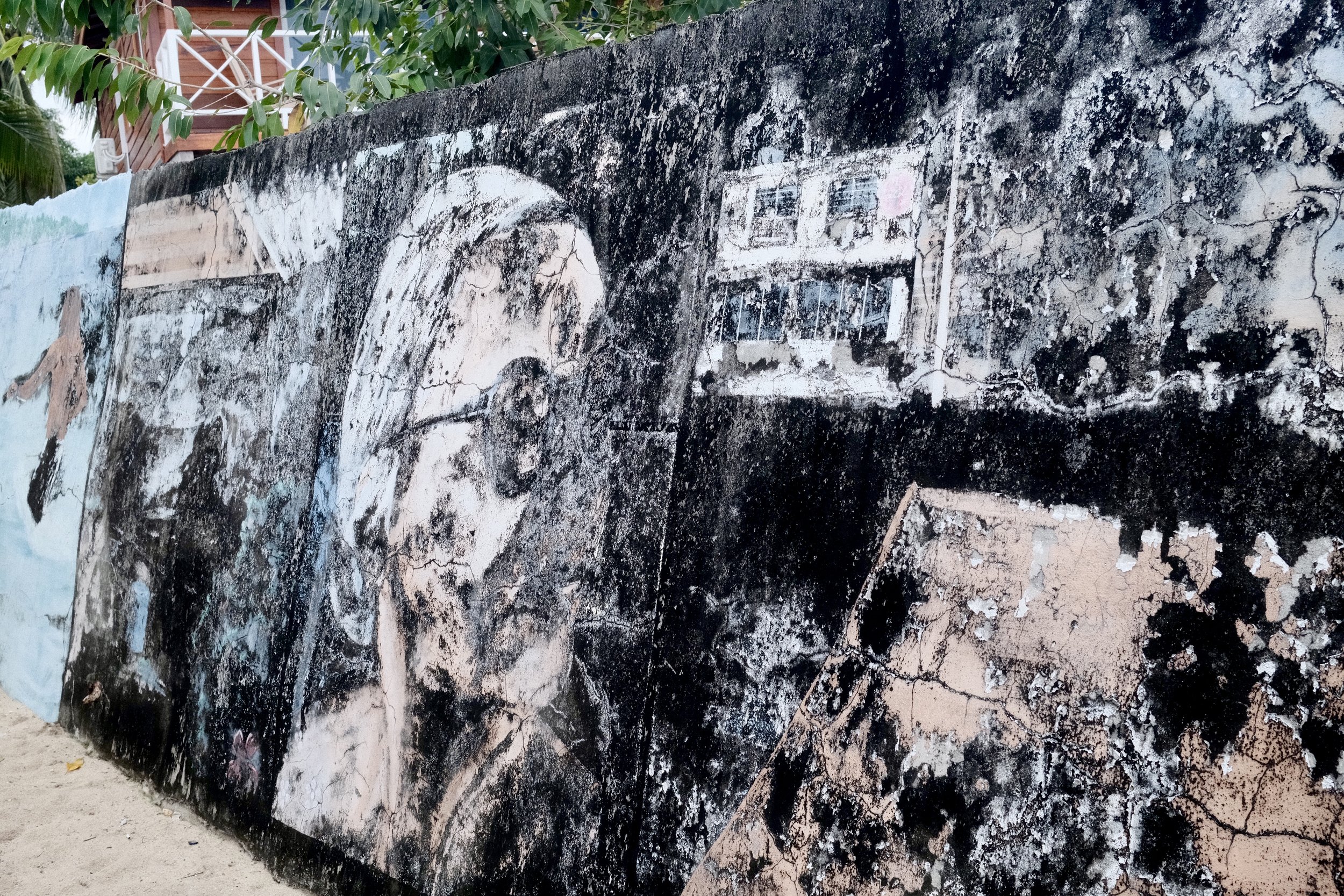 A mural painted on a wall featuring a portrait of a person wearing glasses, surrounded by urban scenes and abstract elements in black, white, and muted tones.