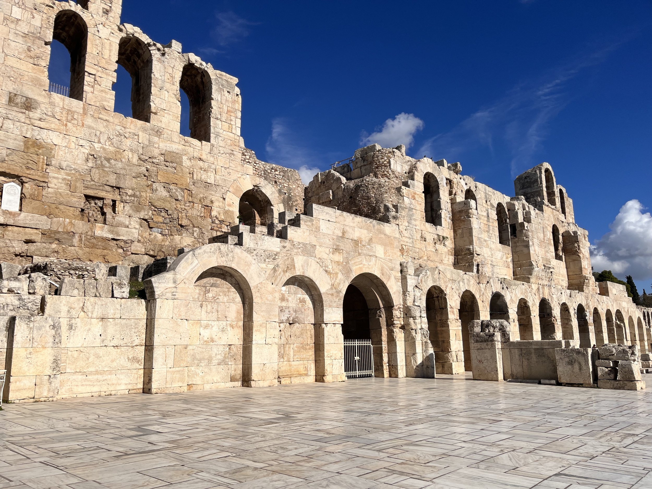 Ancient stone ruins of a large amphitheater or coliseum with tall arches and multiple stories, under a blue sky with a few clouds.