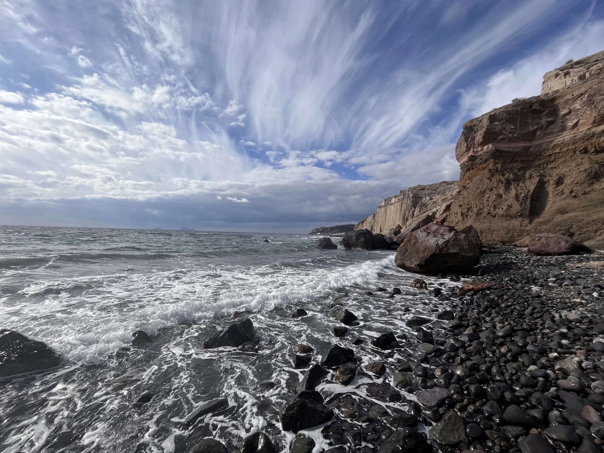 A rocky beach with crashing waves, steep cliffs, and a cloudy sky.