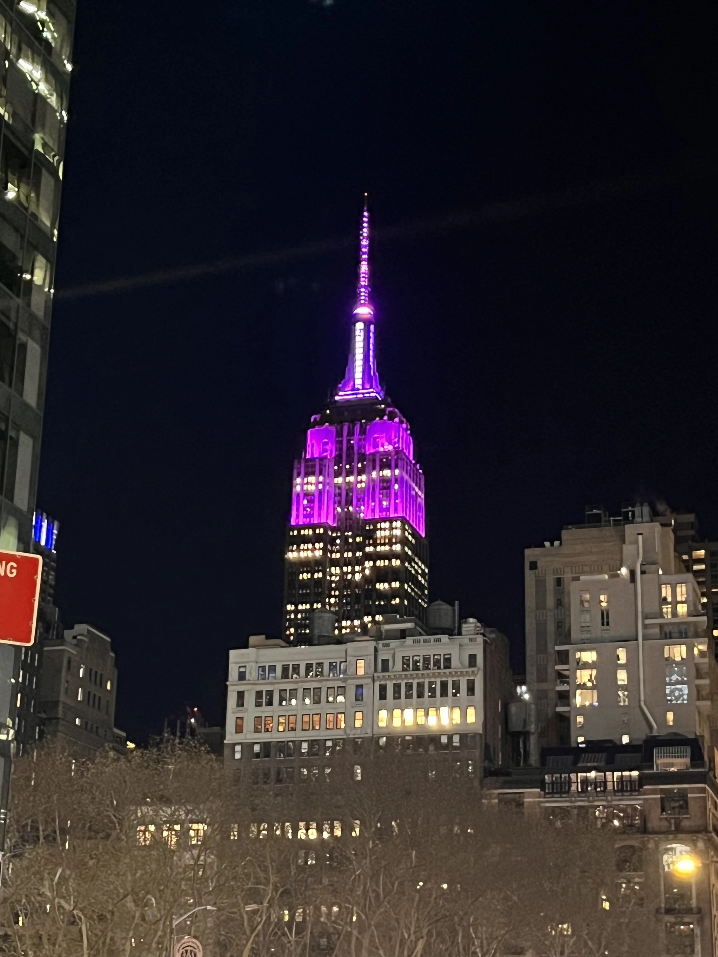 Night view of the Empire State Building illuminated in purple lights in New York City with surrounding buildings.