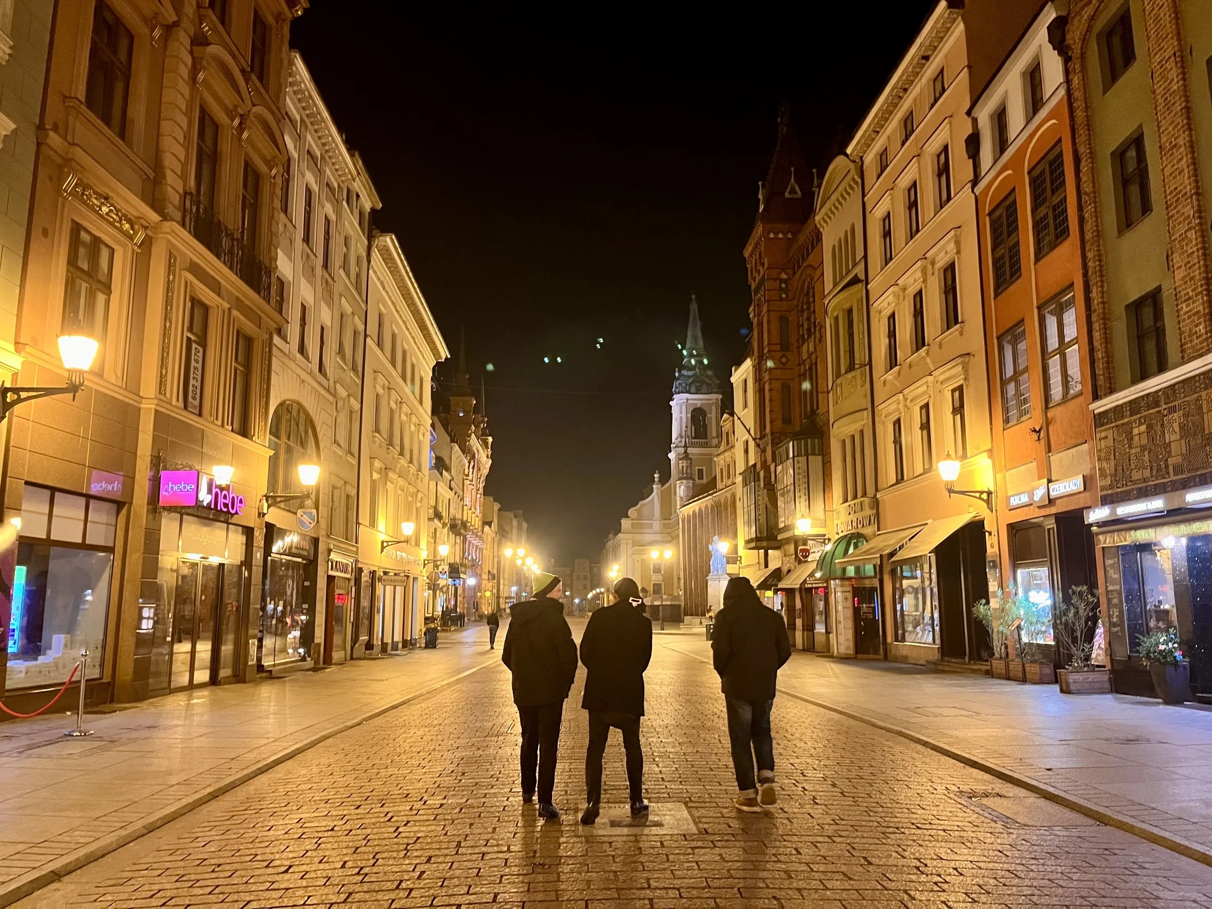 Three people walking along a cobblestone street at night, lined with illuminated buildings, with a church tower visible in the background.