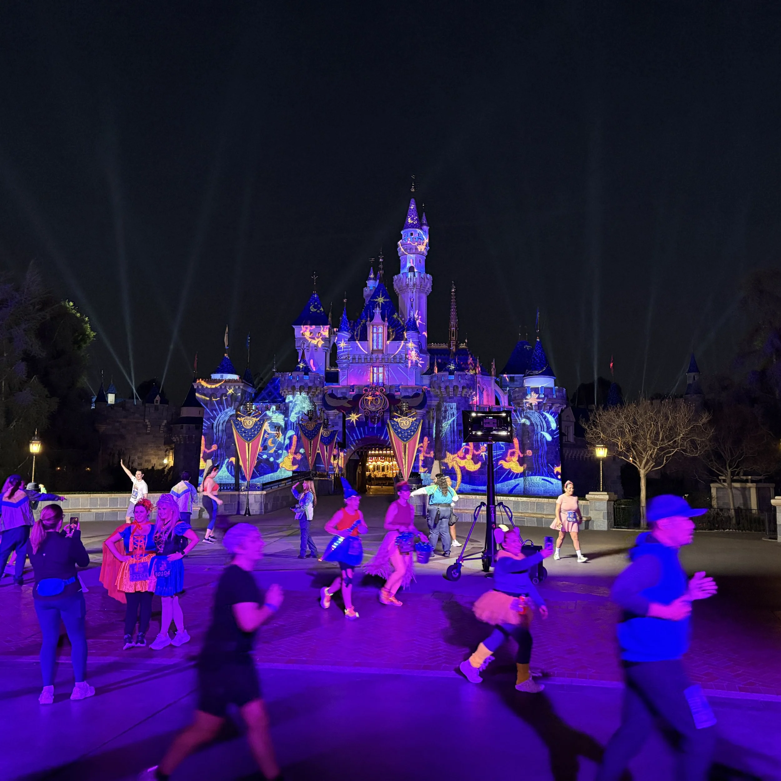 A nighttime scene at a Disney park showing Sleeping Beauty Castle illuminated with colorful lights and projections, with people walking and dancing in front of it.