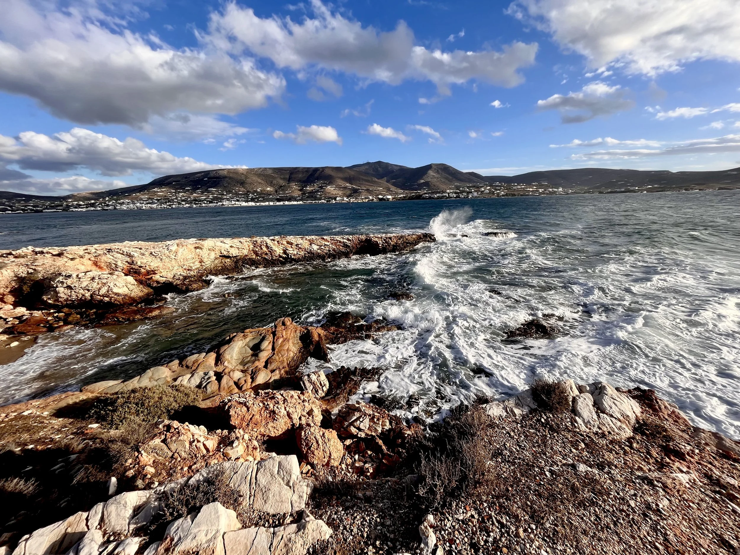 Rocky shoreline with blue water and waves, distant hillside with houses, partly cloudy sky.
