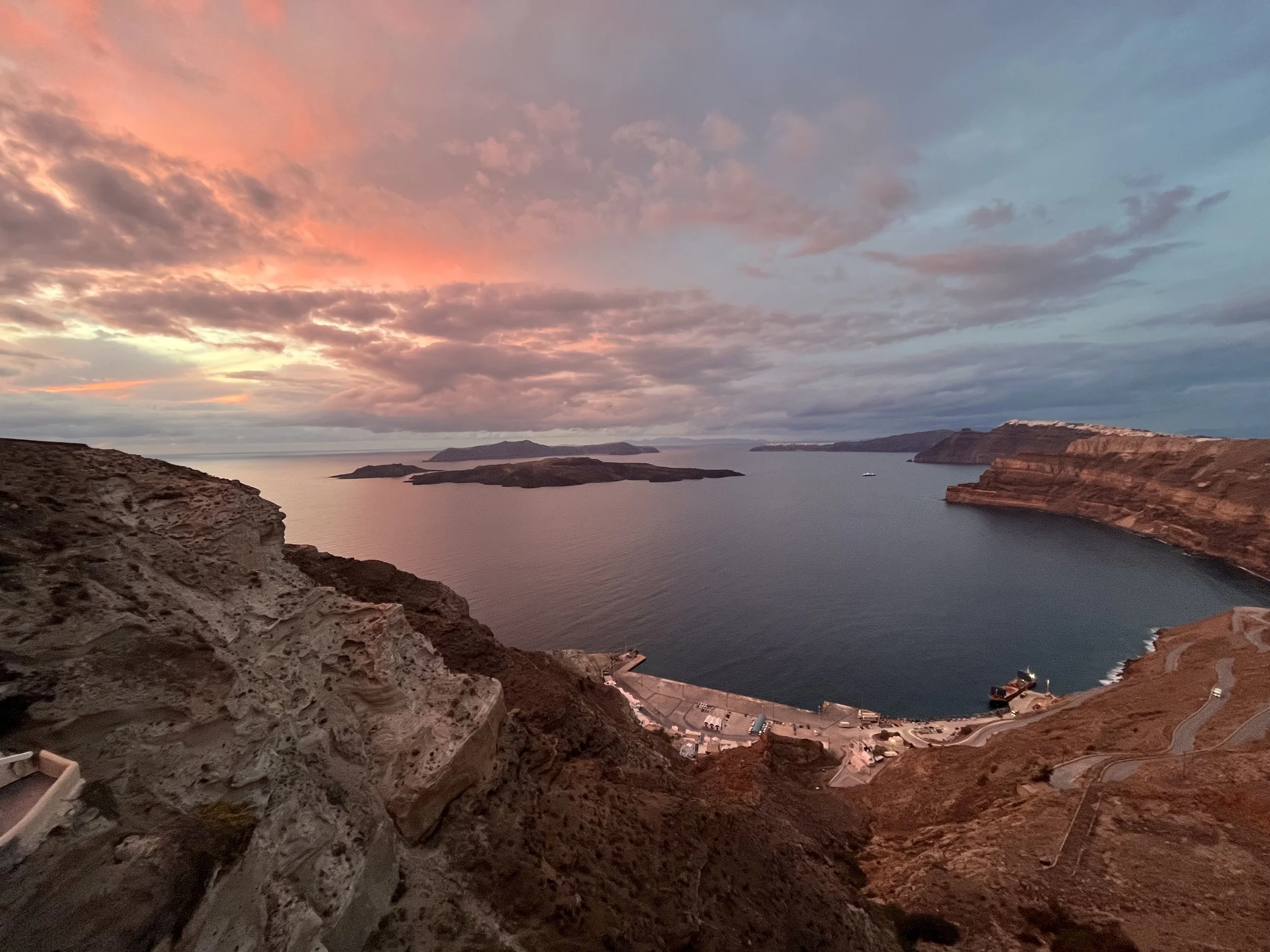 A sunset over the caldera of Santorini with pink and orange clouds, rugged cliffs, and a small harbor with boats.