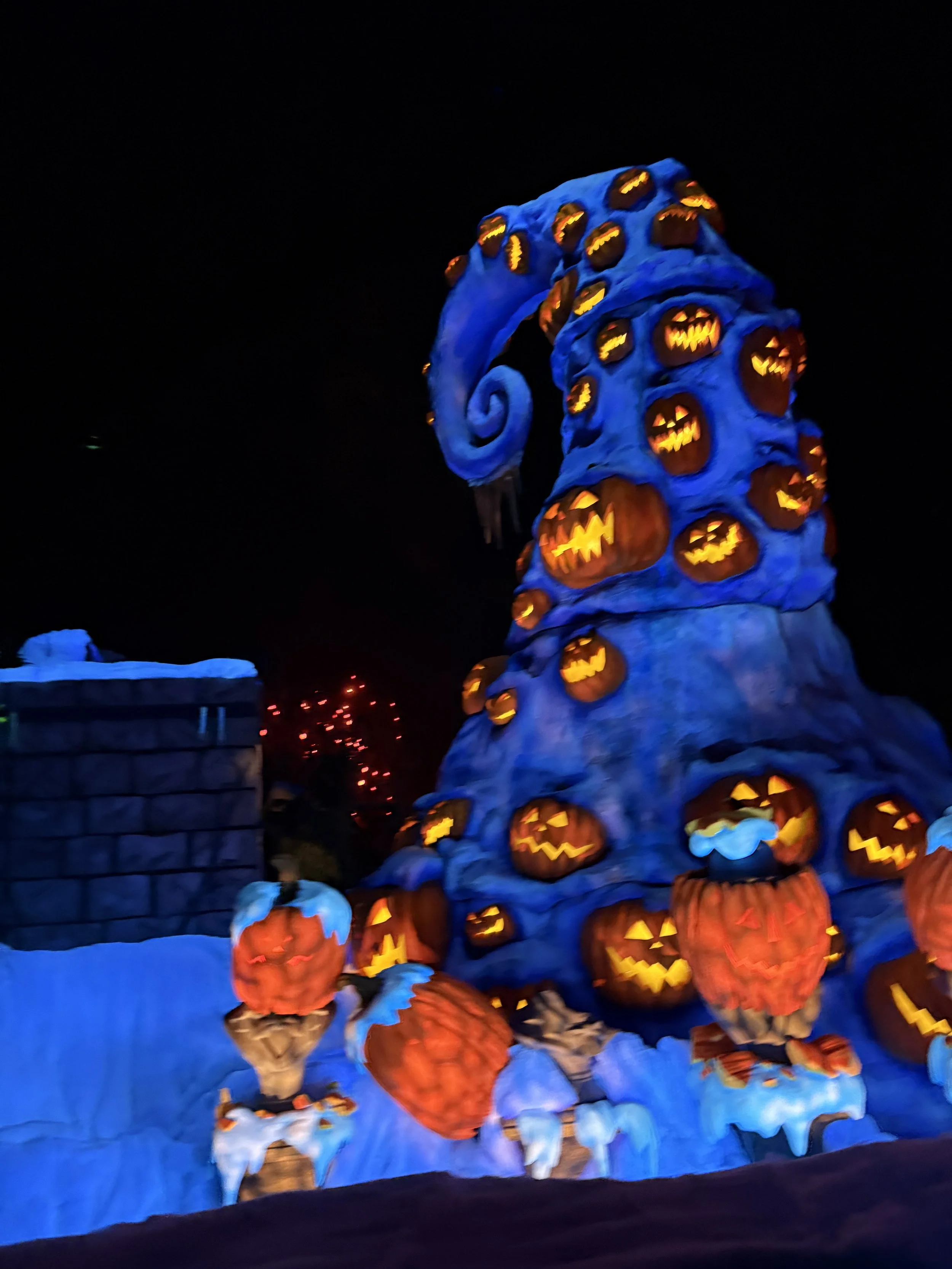 A large illuminated Halloween-themed sculpture with a blue body covered in glowing orange and yellow carved pumpkins and faces, with small figures wearing pumpkin headgear at the base, set against a dark night sky.