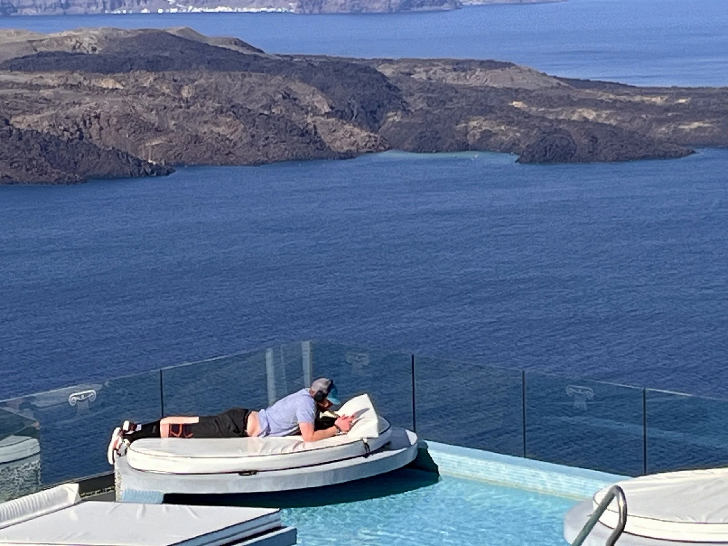 A person lying on a pool float on a deck overlooking a body of water and rocky hills in the background.