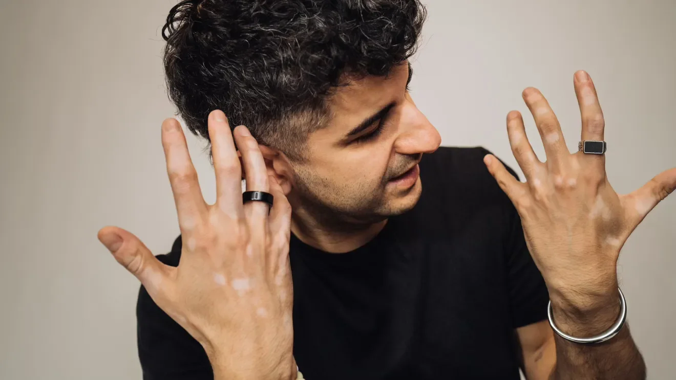Man med svarta kläder, hårlockigt hår, bär ringar och armband, tittar på sina händer.