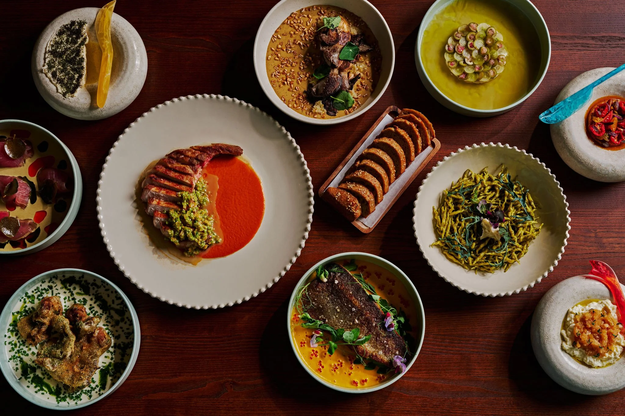 A variety of dishes on a wooden table including sliced steak with sauce, pasta, grilled fish, and several bowls of different culinary items, presented in an elegant, diverse meal setting.