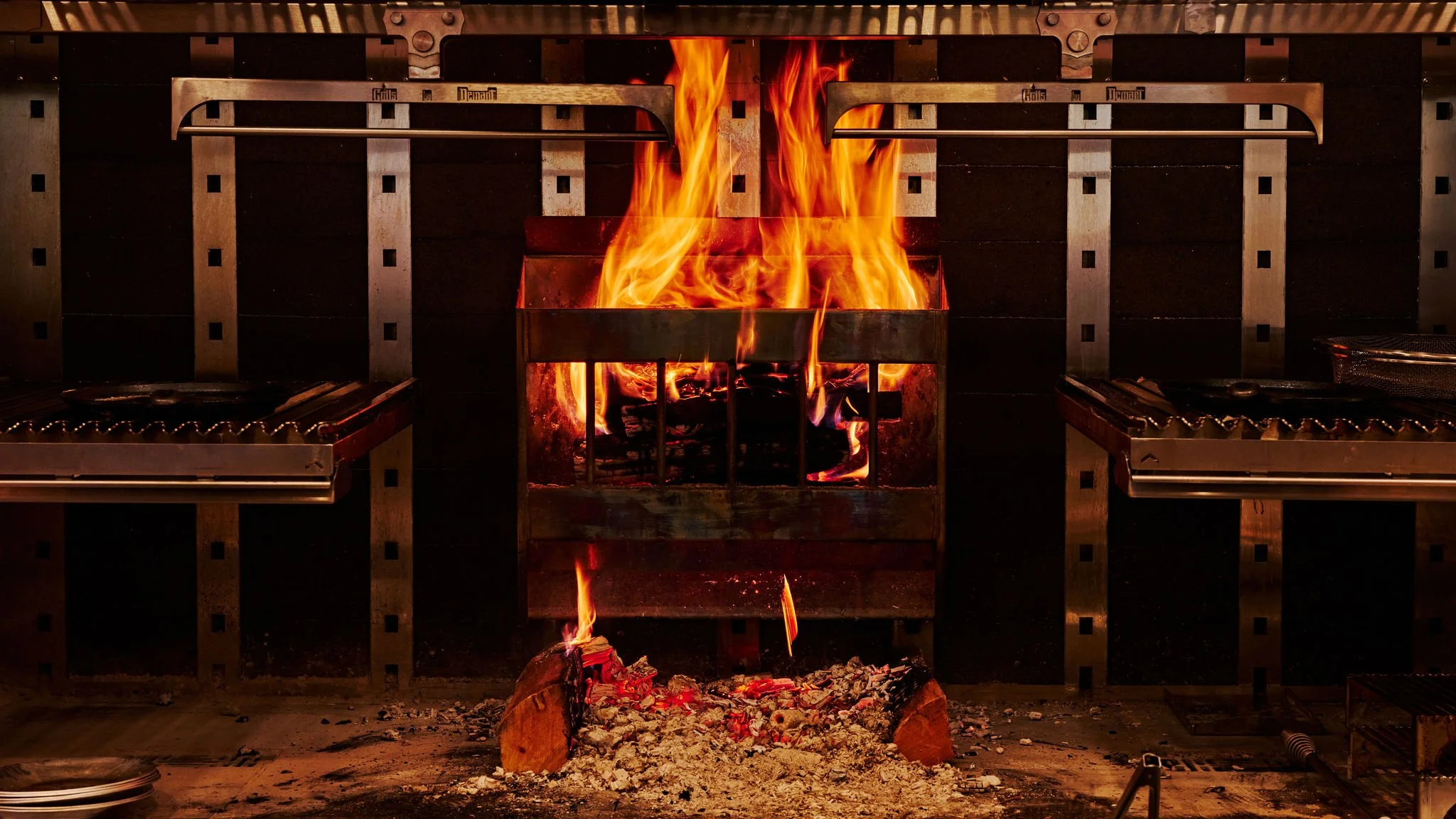 A fire burning in a metal firepit inside a commercial kitchen, with two stainless steel stoves on either side.