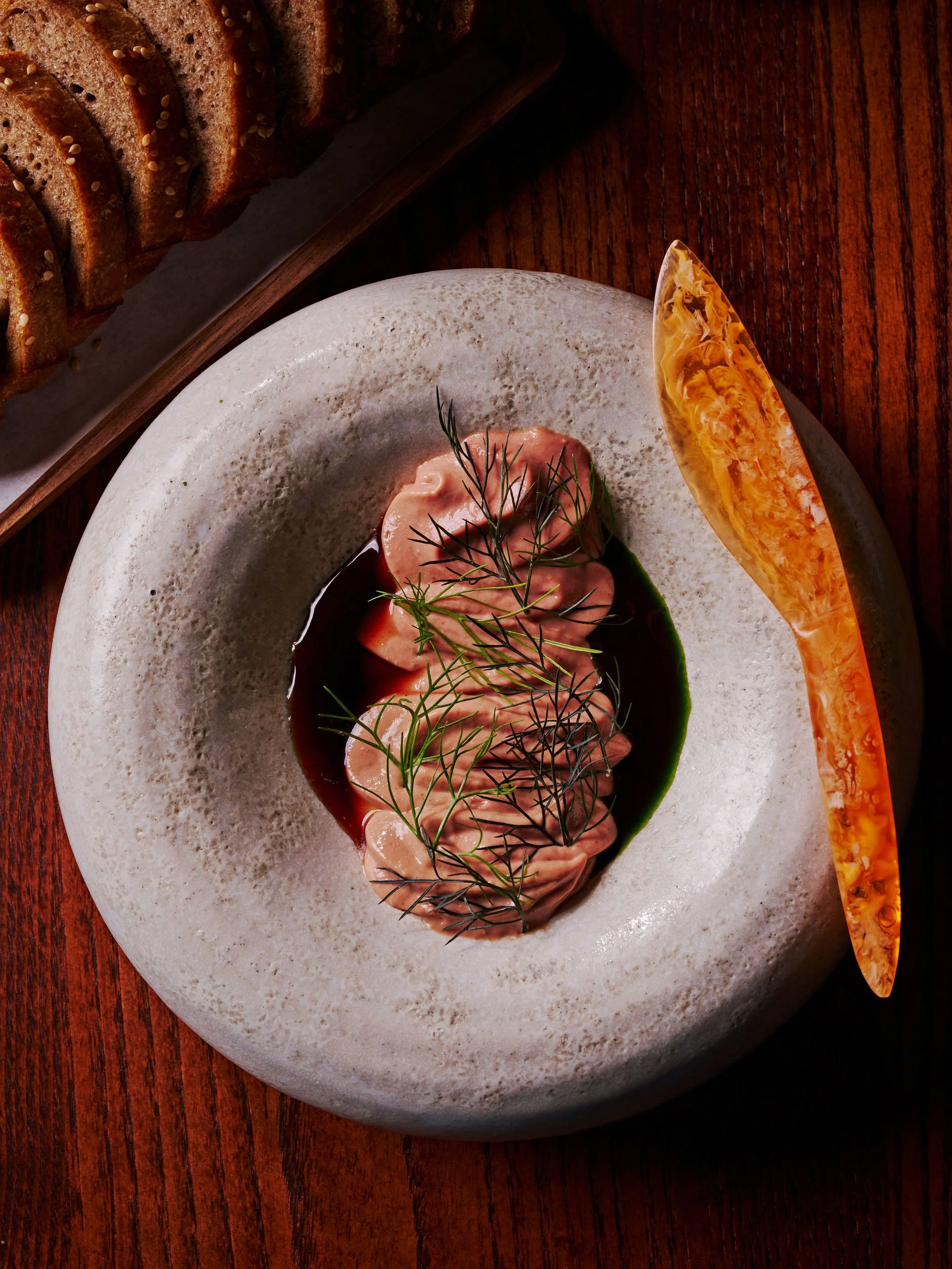 Sliced raw fish, possibly salmon, garnished with fresh dill, served on a white stone plate with a dark sauce and a decorative orange and yellow glass piece. Slices of toasted bread are in the background.