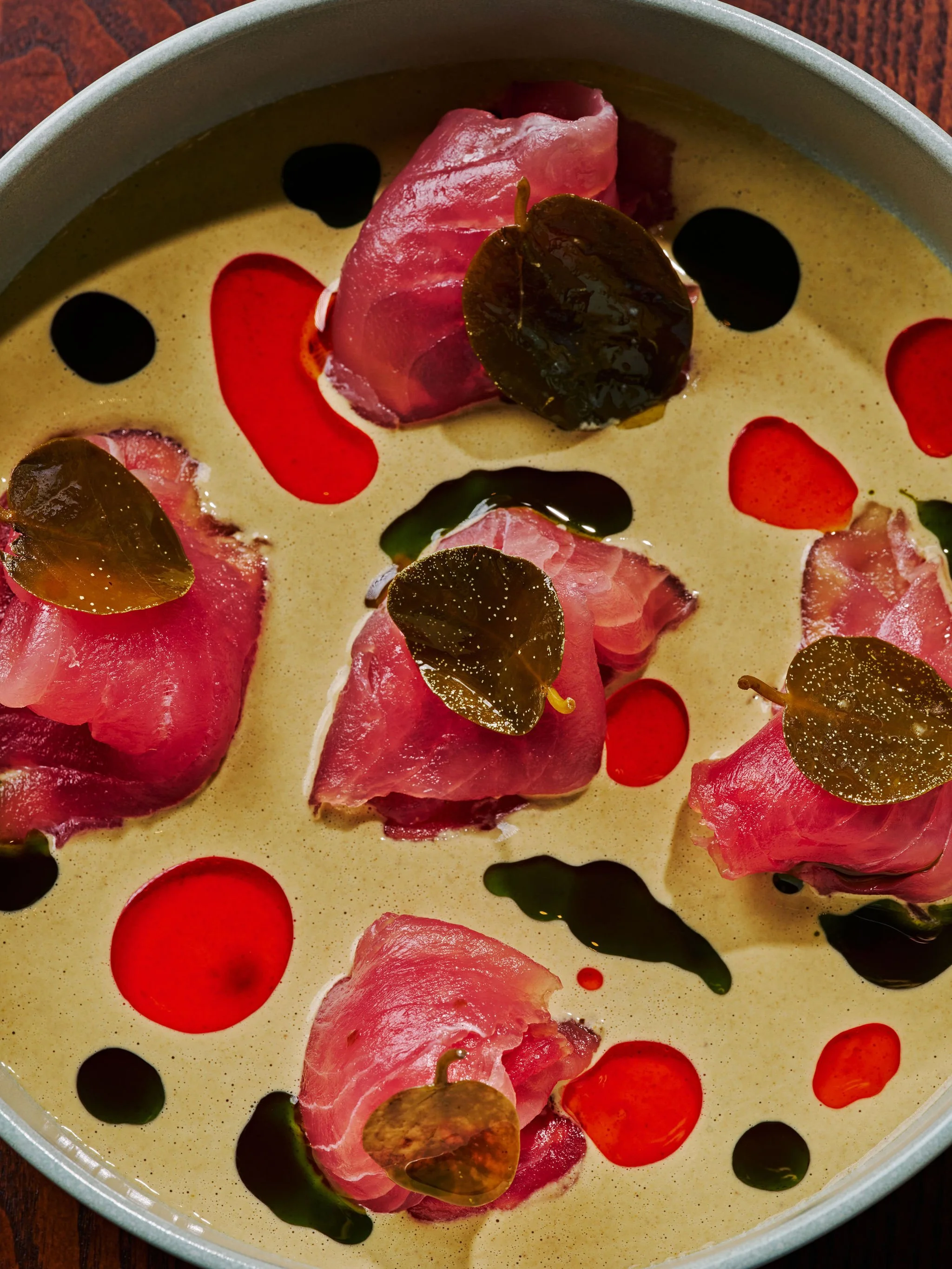 Close-up of a dish with slices of cured meat topped with basil leaves, in a creamy yellow sauce with red and black dots of sauce or oil.