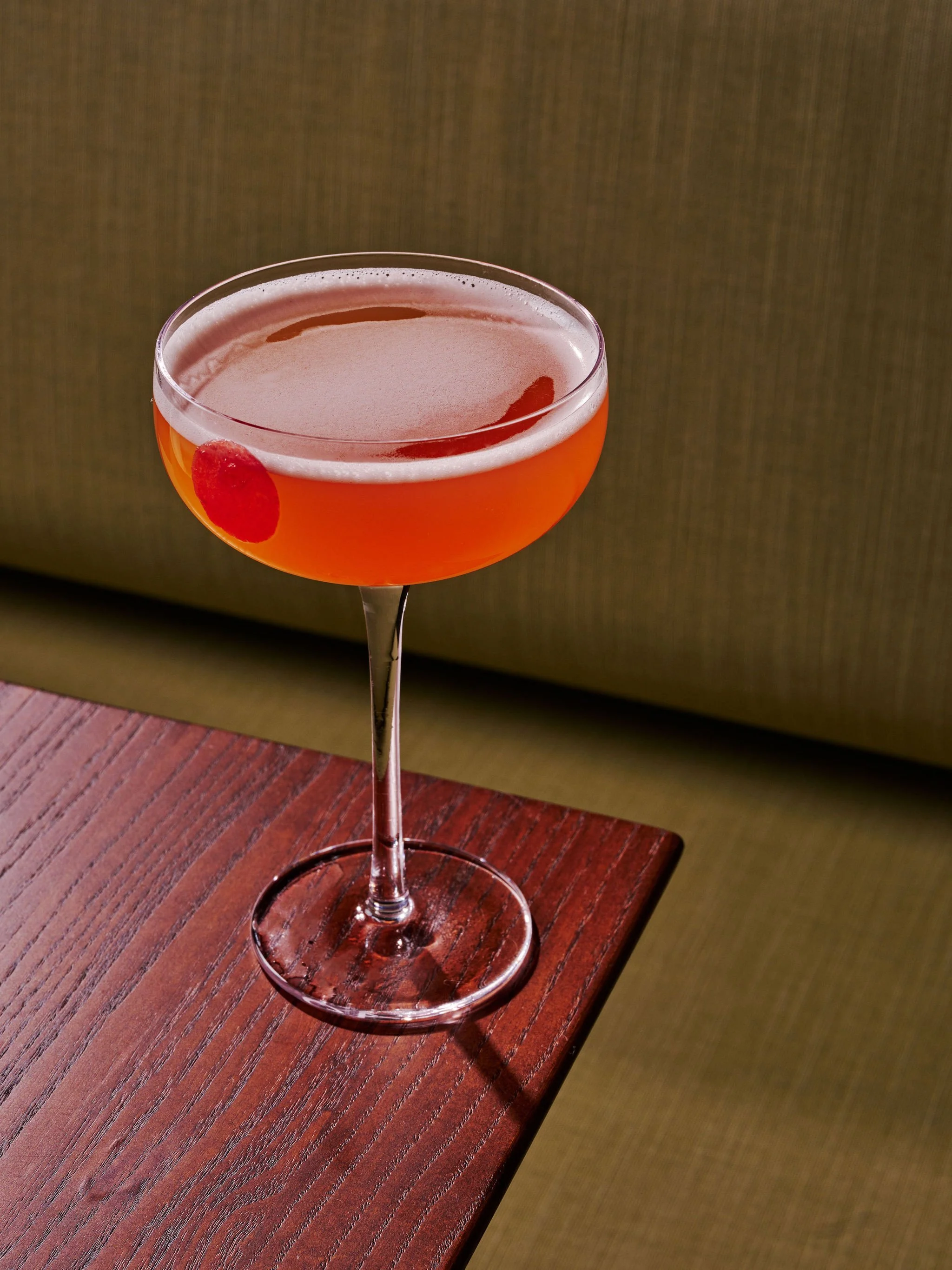 A cocktail glass with an orange-colored drink and a red cherry garnish, placed on a wooden surface.