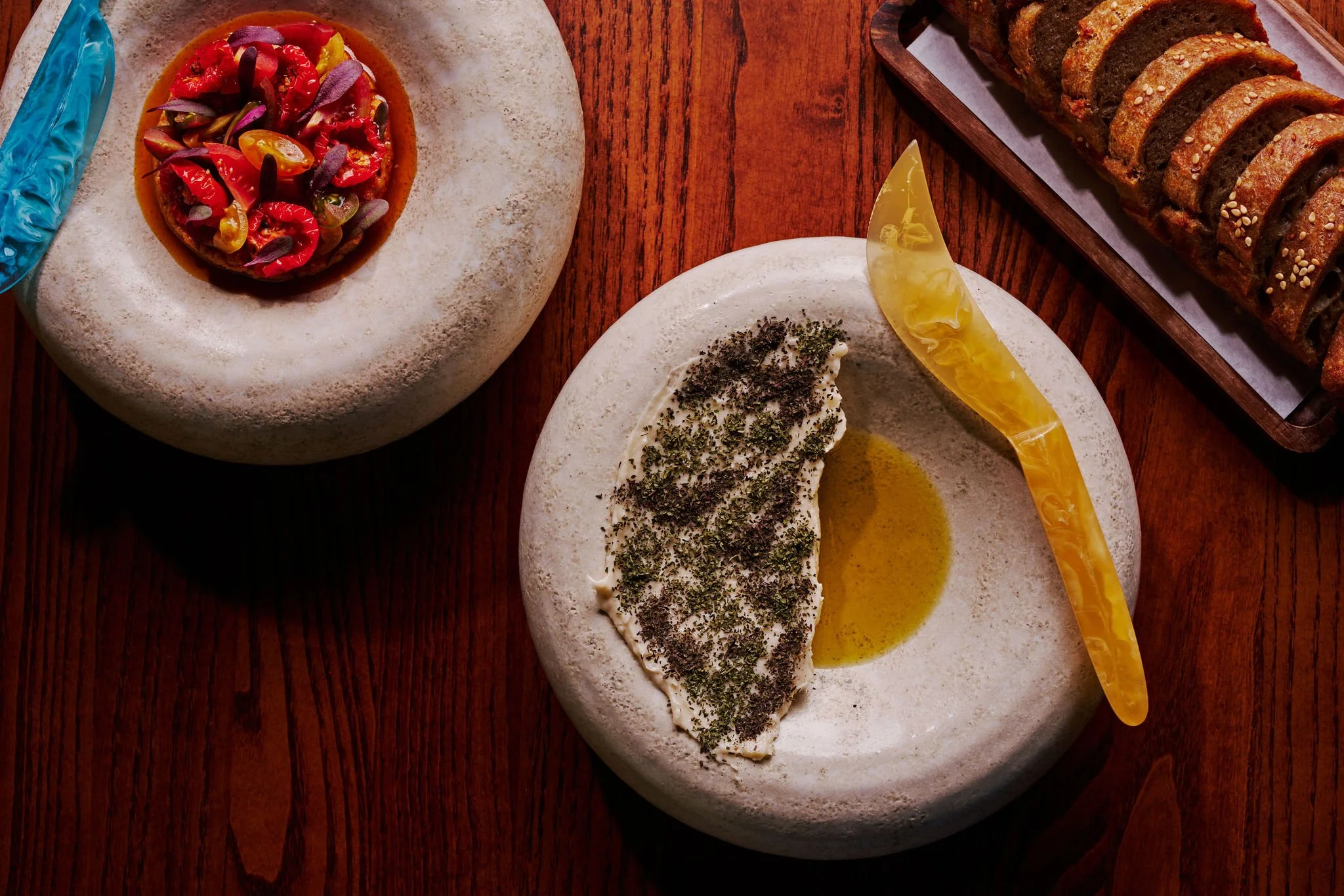 A table with three different dishes: a tomato salad with cherry tomatoes and sliced vegetables served on a white stone plate, a spread of white cheese topped with herbs and a lemon wedge on a white stone plate, and a sliced loaf of bread with sesame 