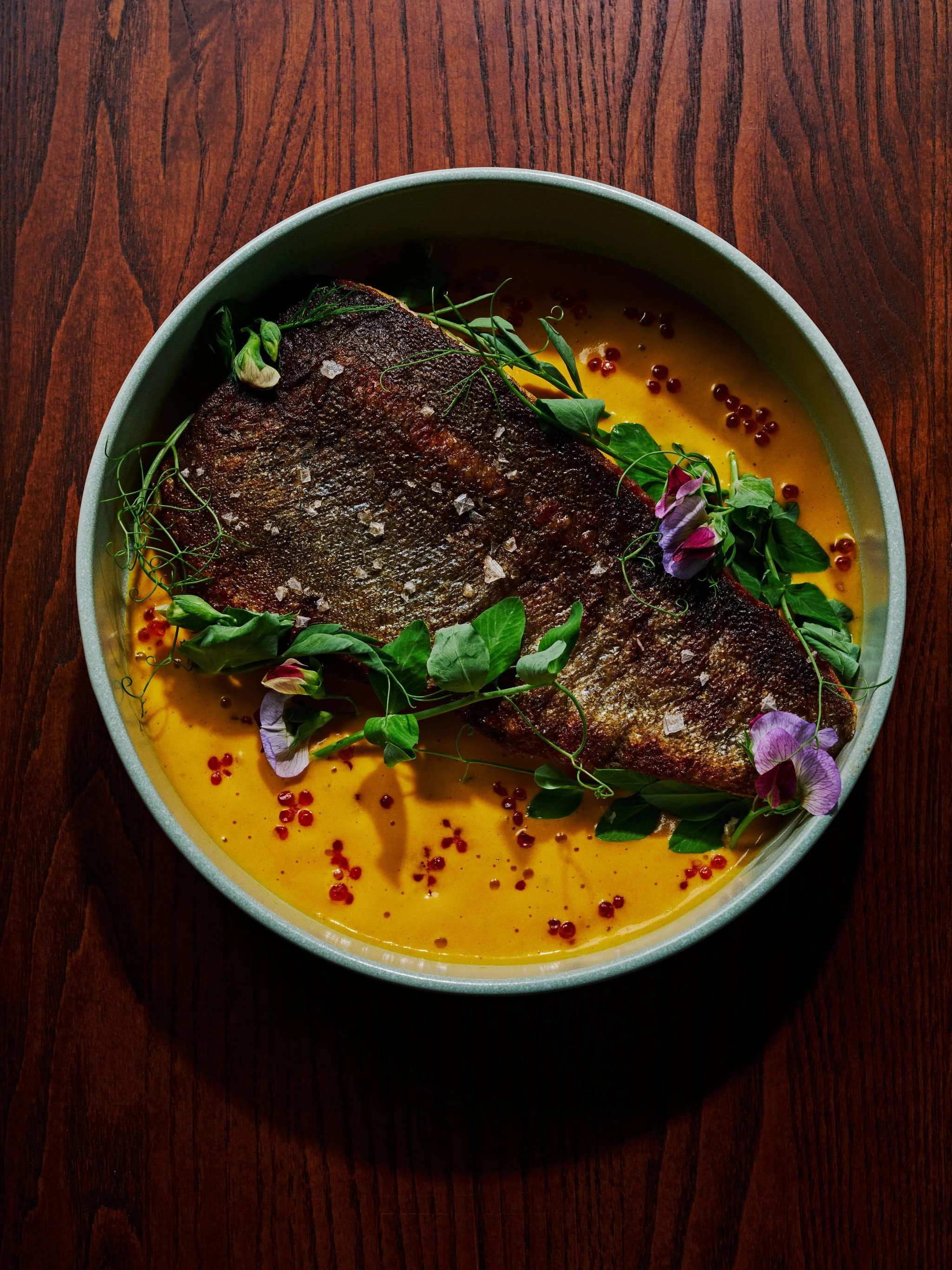 A cooked fish fillet garnished with fresh herbs and edible flowers, served in a bowl with a yellow sauce and red peppercorns, on a wooden table.