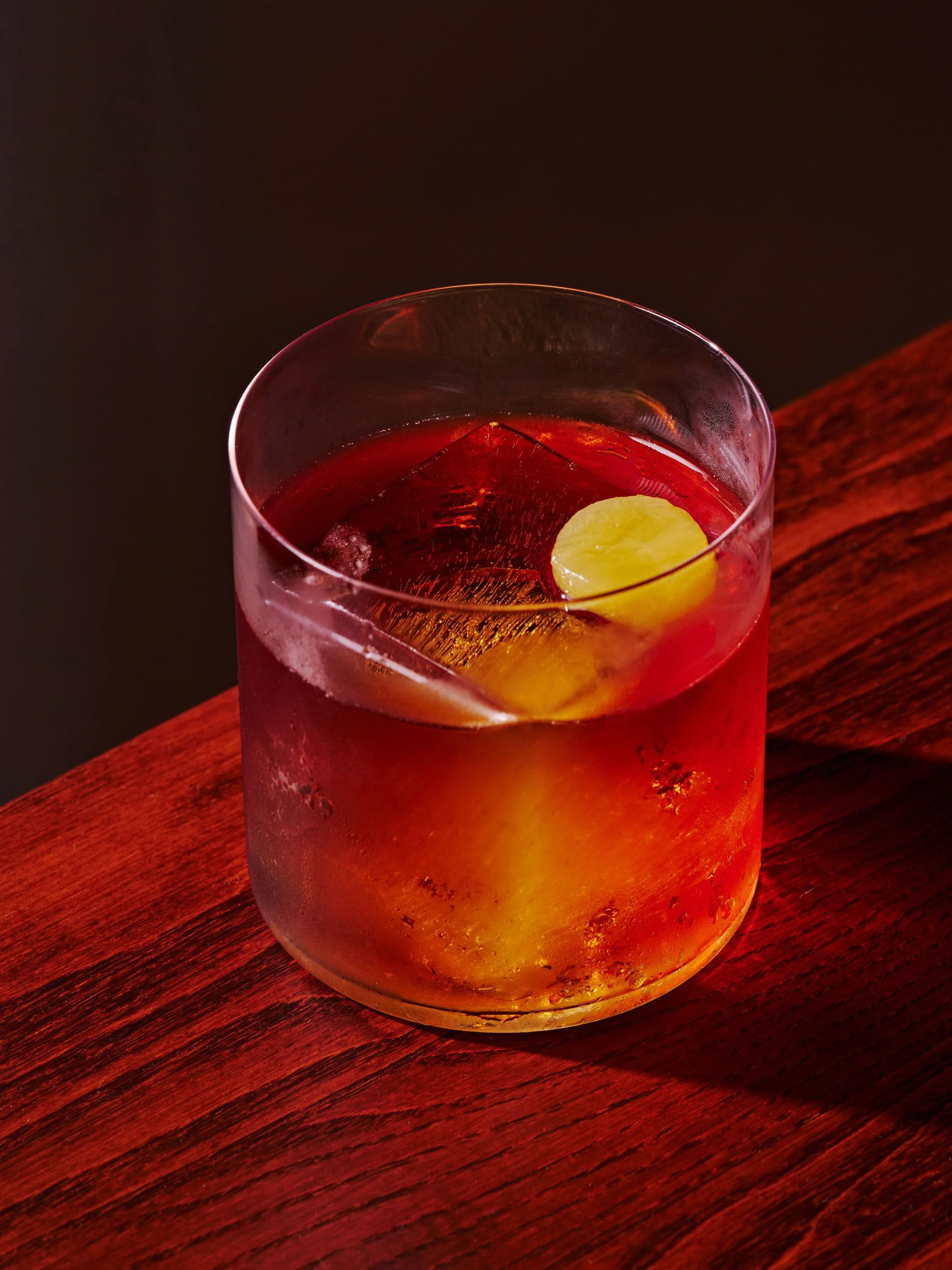 A glass of red-colored cocktail with lemon slices on a wooden surface.