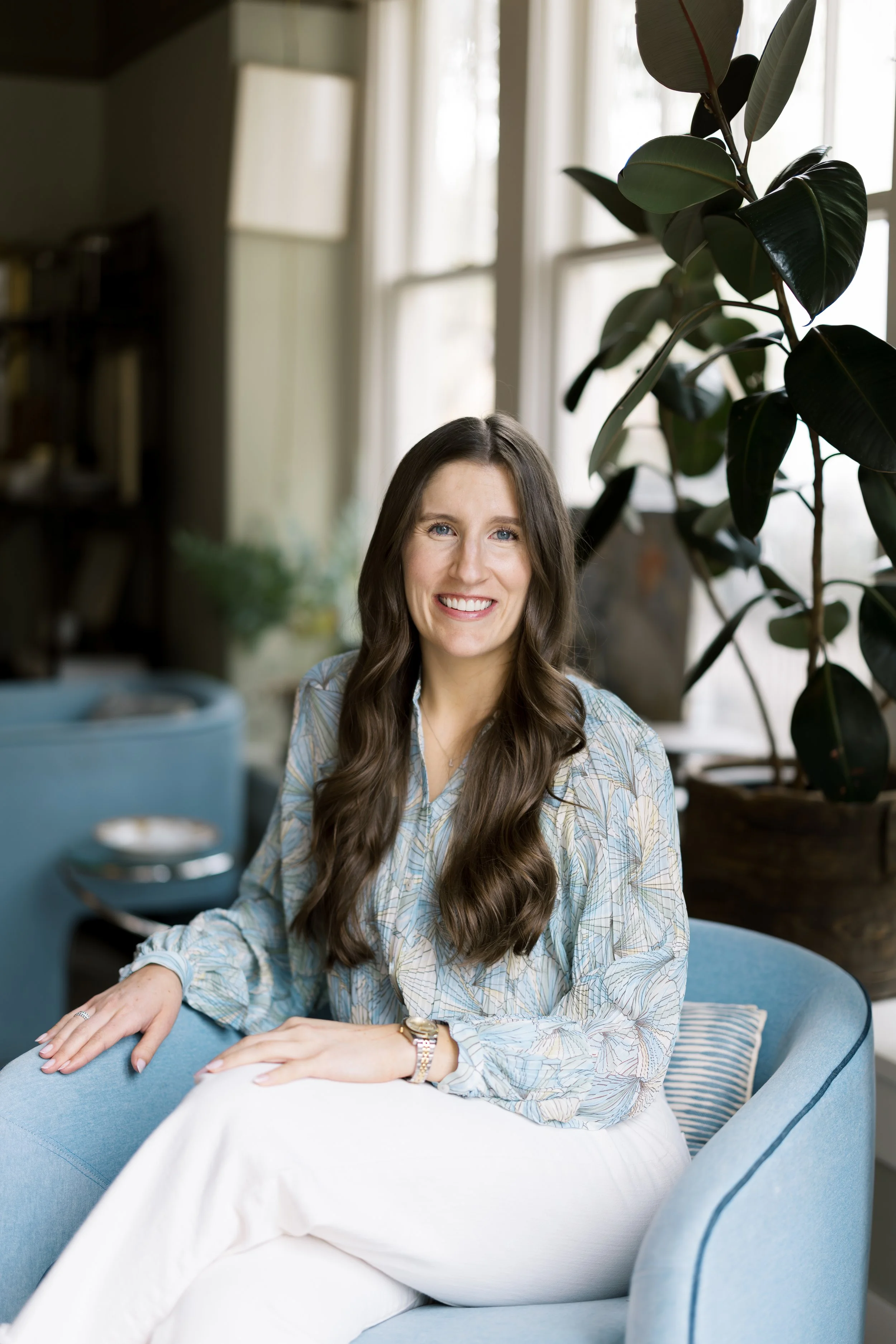 A woman with long brown hair and blue eyes sits on a blue sofa in a cozy, well-lit room with large windows, a green potted plant, and a bookshelf in the background, smiling at the camera.