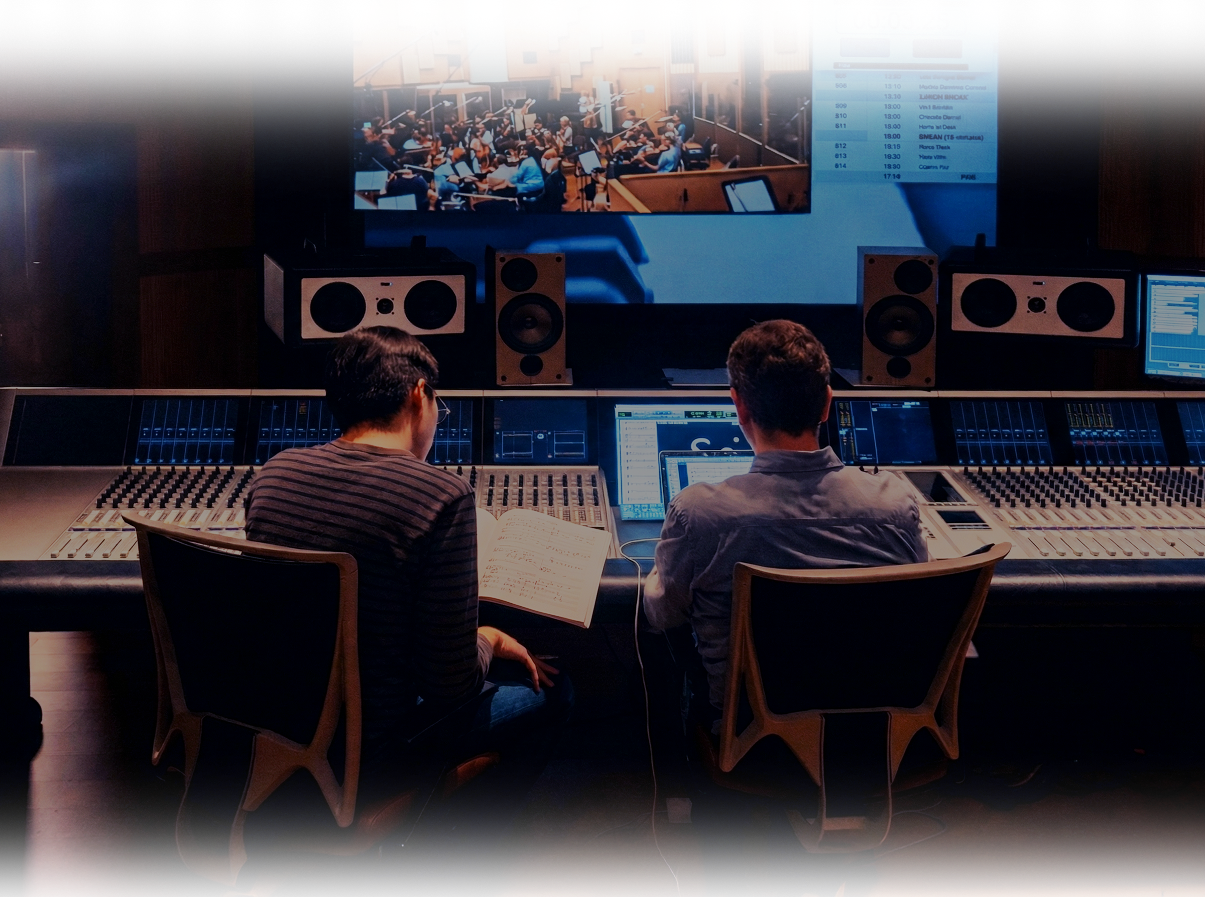 음악 스튜디오에서 두 명의 남성이 작업하는 모습, 큰 오디오 믹서와 스피커, 모니터가 설치되어 있고, 큰 화면에 오케스트라 영상이 보여지는 모습