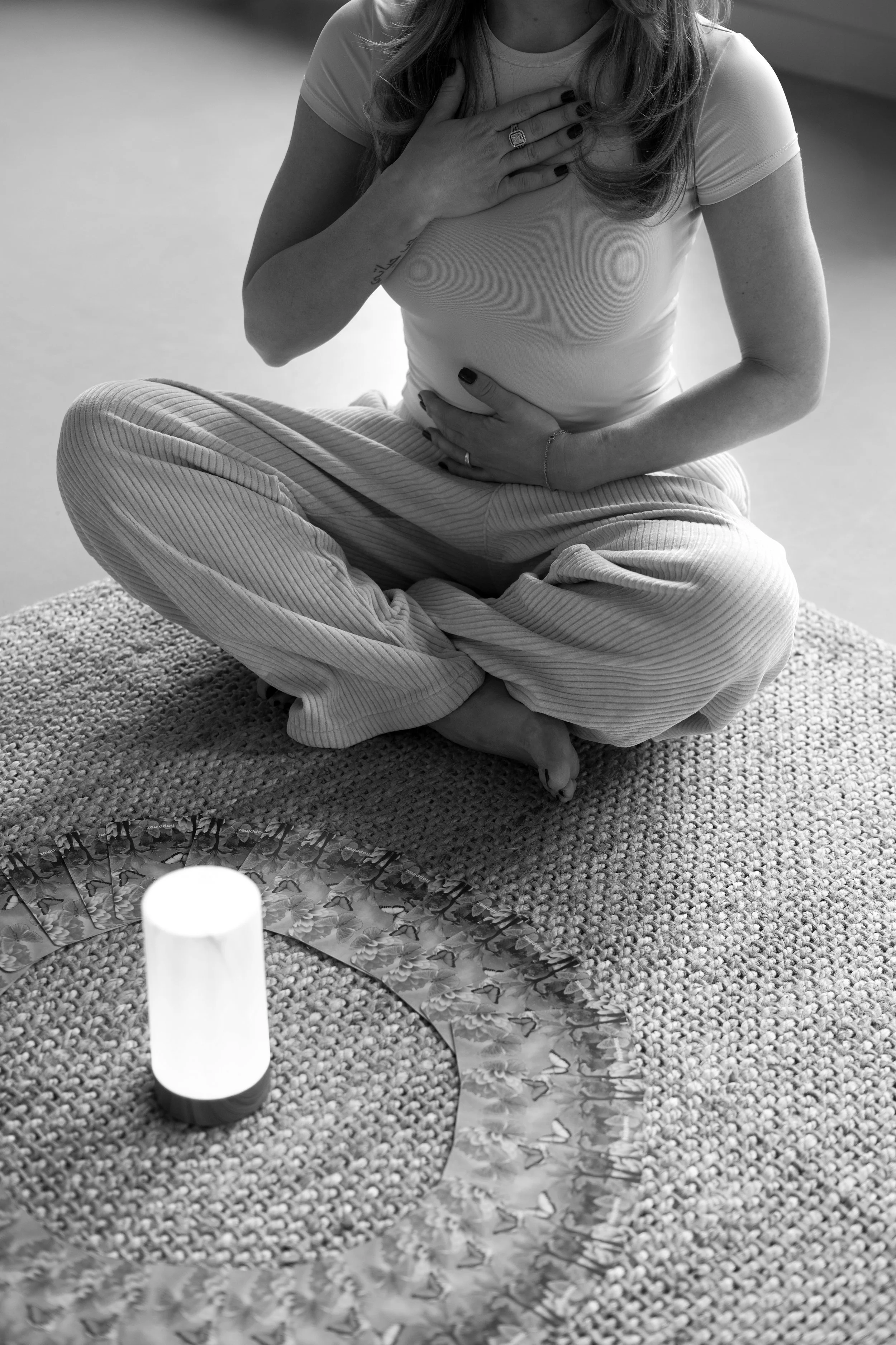 A woman sitting cross-legged on a textured rug, holding her chest and stomach, with a glowing candle on a decorated surface in front of her.