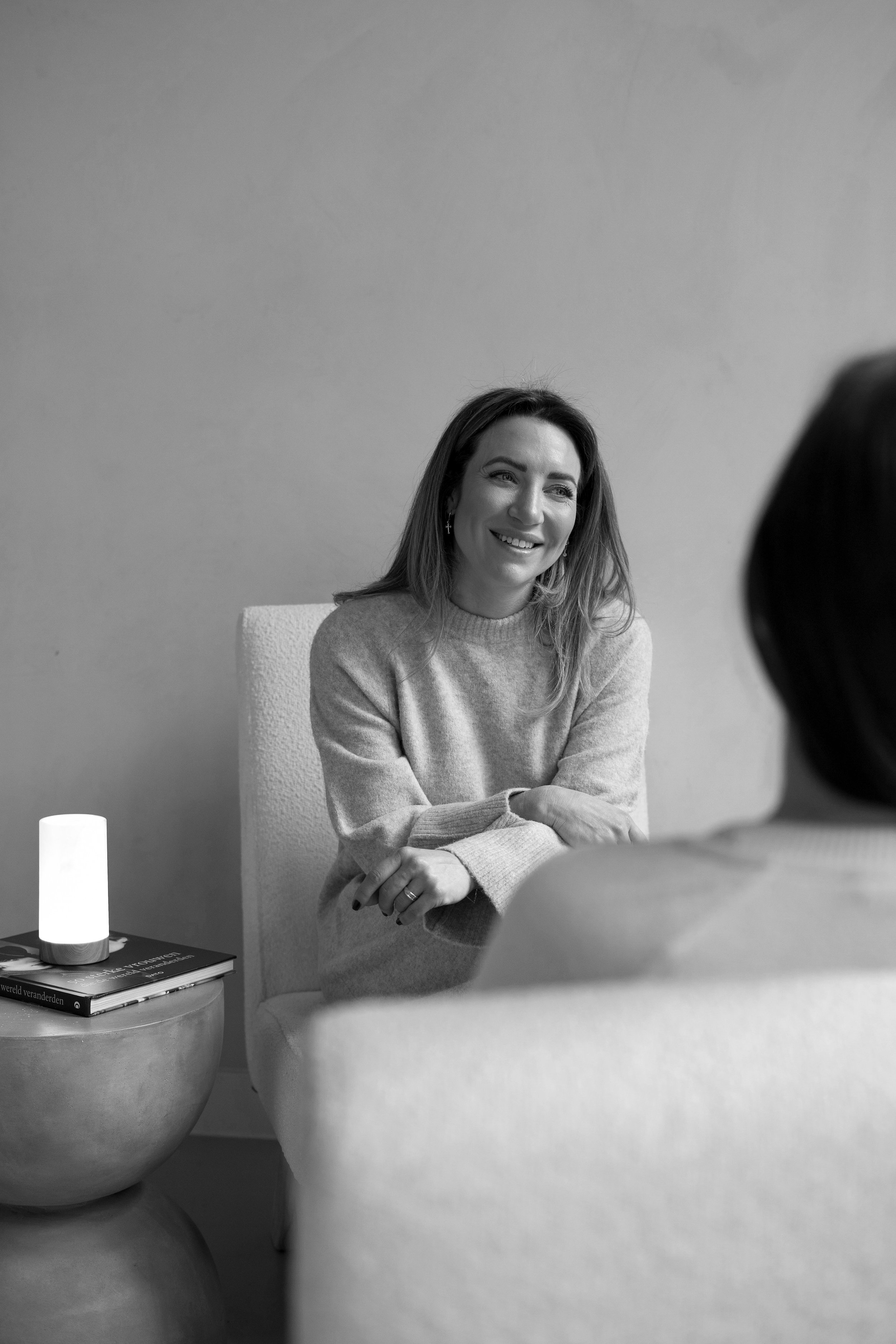 A woman smiling while sitting in a chair during a conversation, with another person partially visible in the foreground, a small table with a book and a lamp next to her.