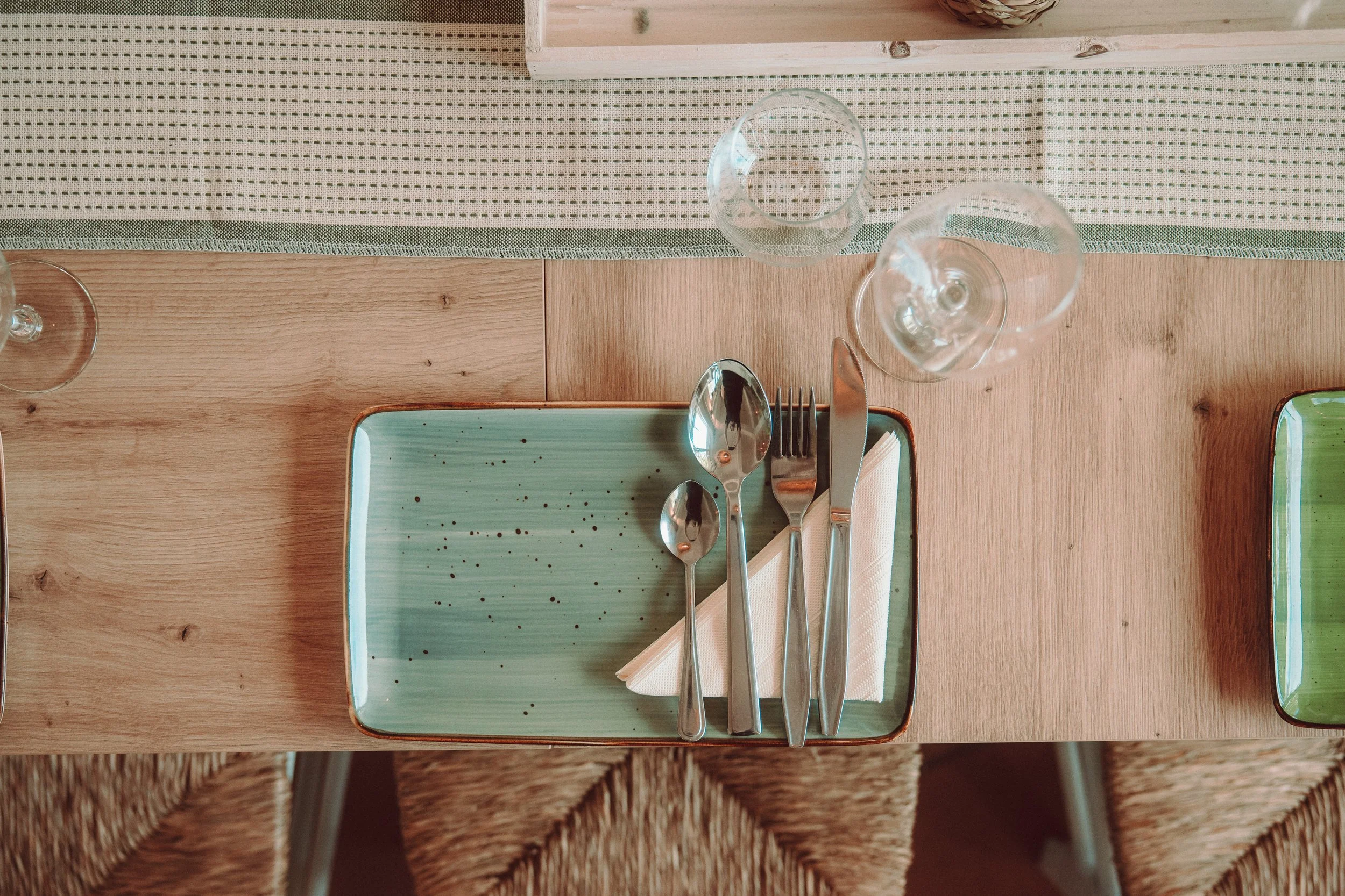 Ein gedeckter Esstisch mit Besteck, einem grünen Teller, Weingläsern und Trinkgläsern, in einer angenehmen, natürlich beleuchteten Umgebung.