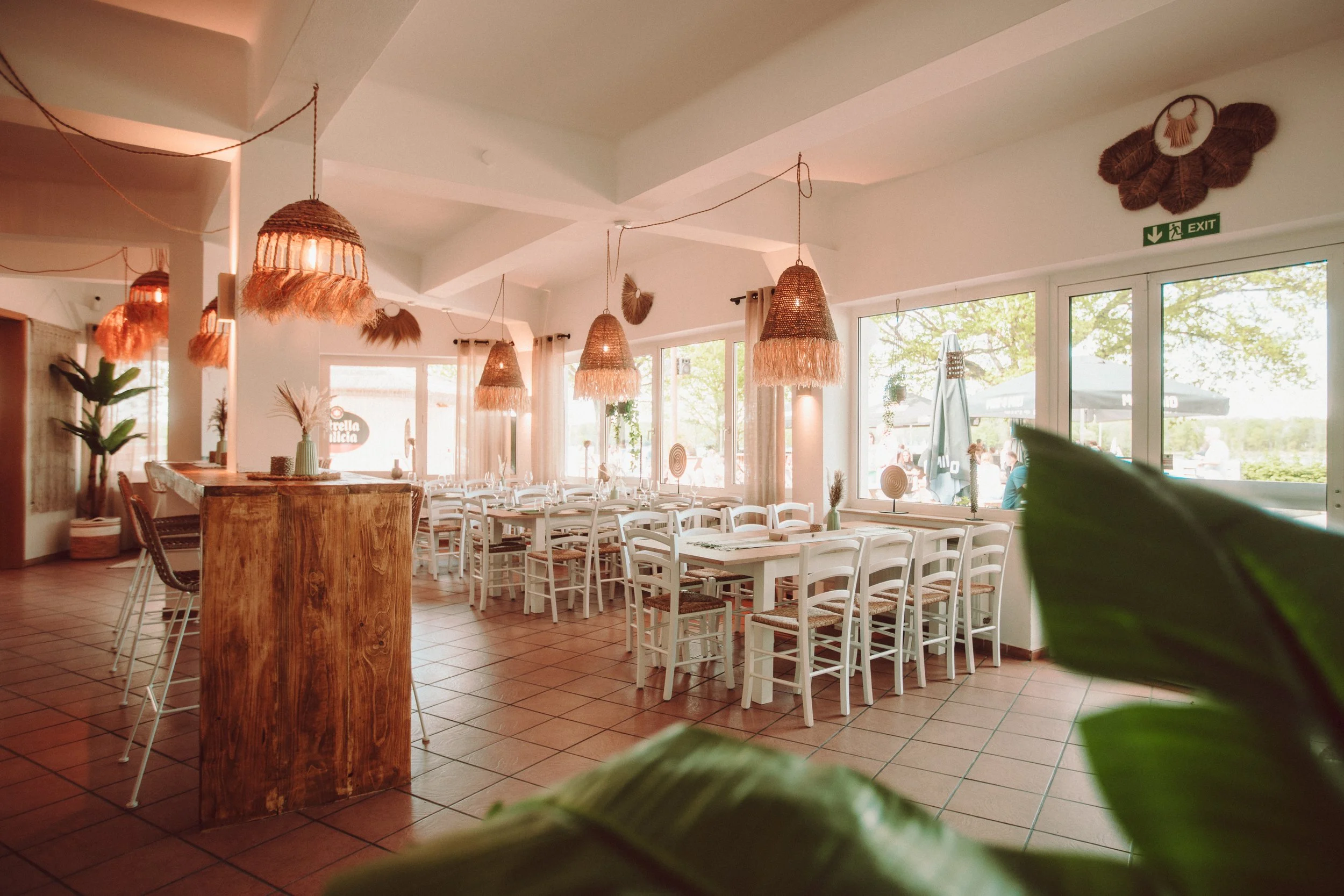 Der Veranstaltungsraum der Sunset Beach Bar, ausgestattet mit weißen Stühlen, Holztischen und geflochtenen Lampenschirmen, große Fenster mit Blick auf den Außenbereich.