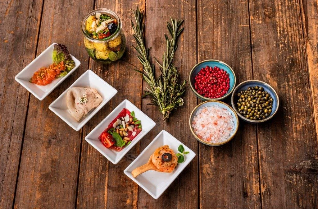 Verschiedene mediterrane Gewürze, Kräuter und eingelegte Lebensmittel auf einem Holztisch.