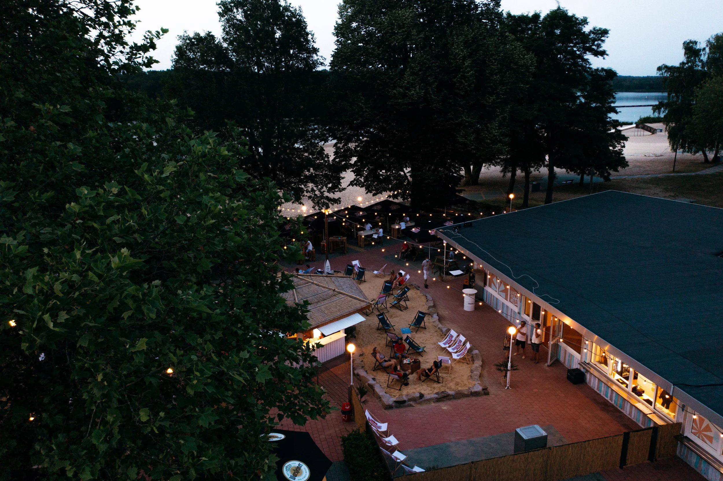 Dämmerungsaufnahme des Außenbereichs der Sunset Beach Bar mit Sitzgelegenheiten, Lichterketten und einigen Gästen, umgeben von Bäumen neben dem See in Haltern.
