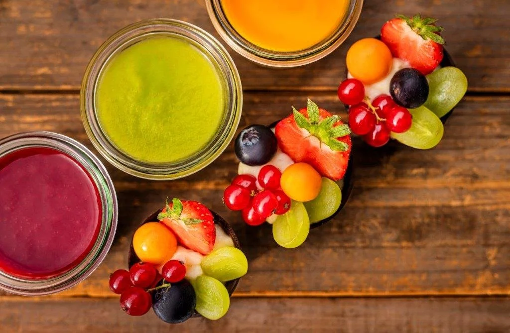 Drei Gläser mit bunten Fruchtsäften und mehrere mit exotischen Früchten wie Erdbeeren, Weintrauben, und Passionsfrucht, auf einer Holztisch.