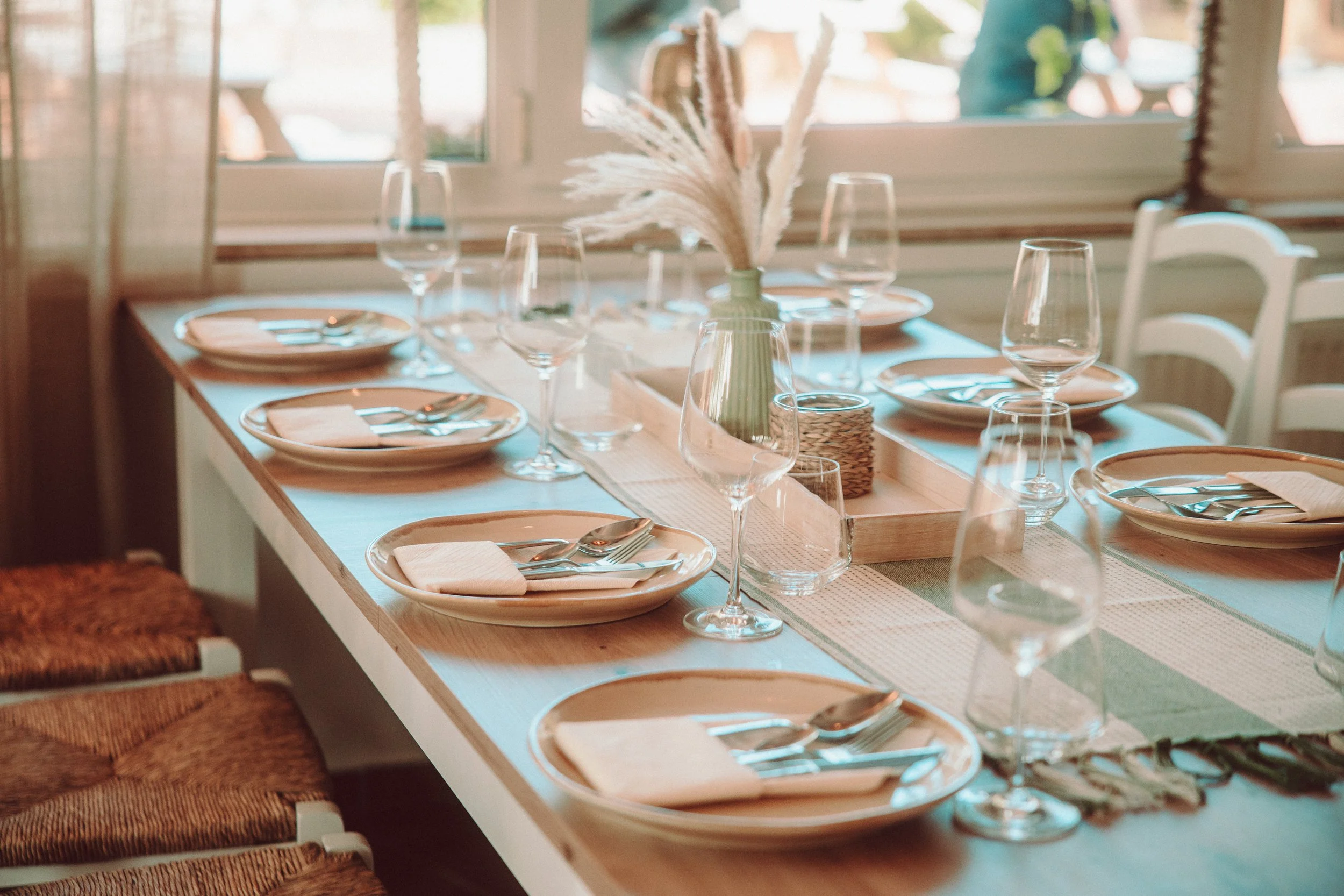 Ein schön gedeckter Esstisch mit weißen Tellern, Gläsern und Besteck, dekoriert mit einer Vase mit Trockenblumen, vor einem Fenster in einer gemütlichen, hellen Umgebung.