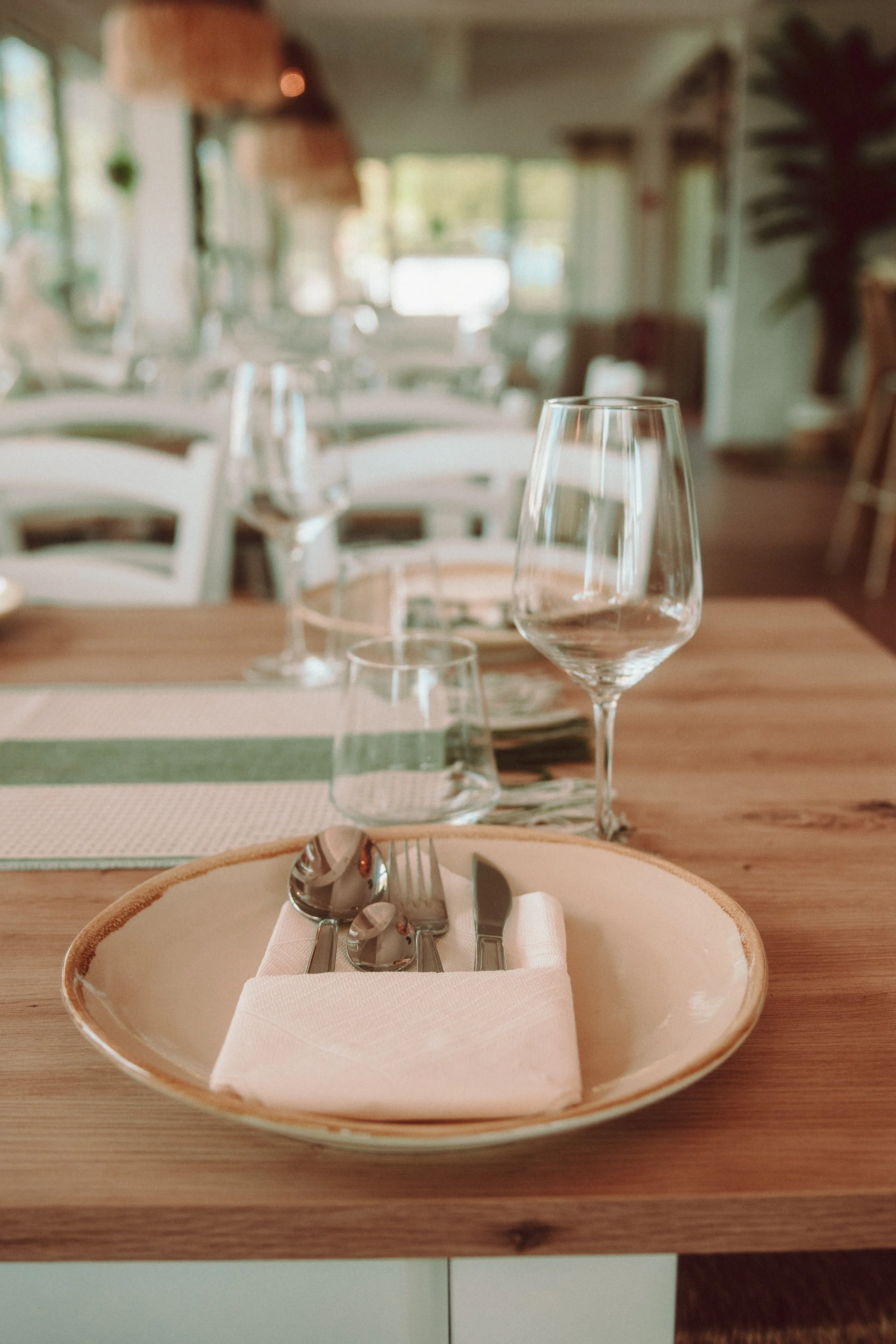 Ein gedeckter Esstisch mit Wein- und Wassergläser, Besteck auf einer weißen Serviette auf einem keramischen Teller, in einem gemütlichen Restaurant mit natürlichem Licht.