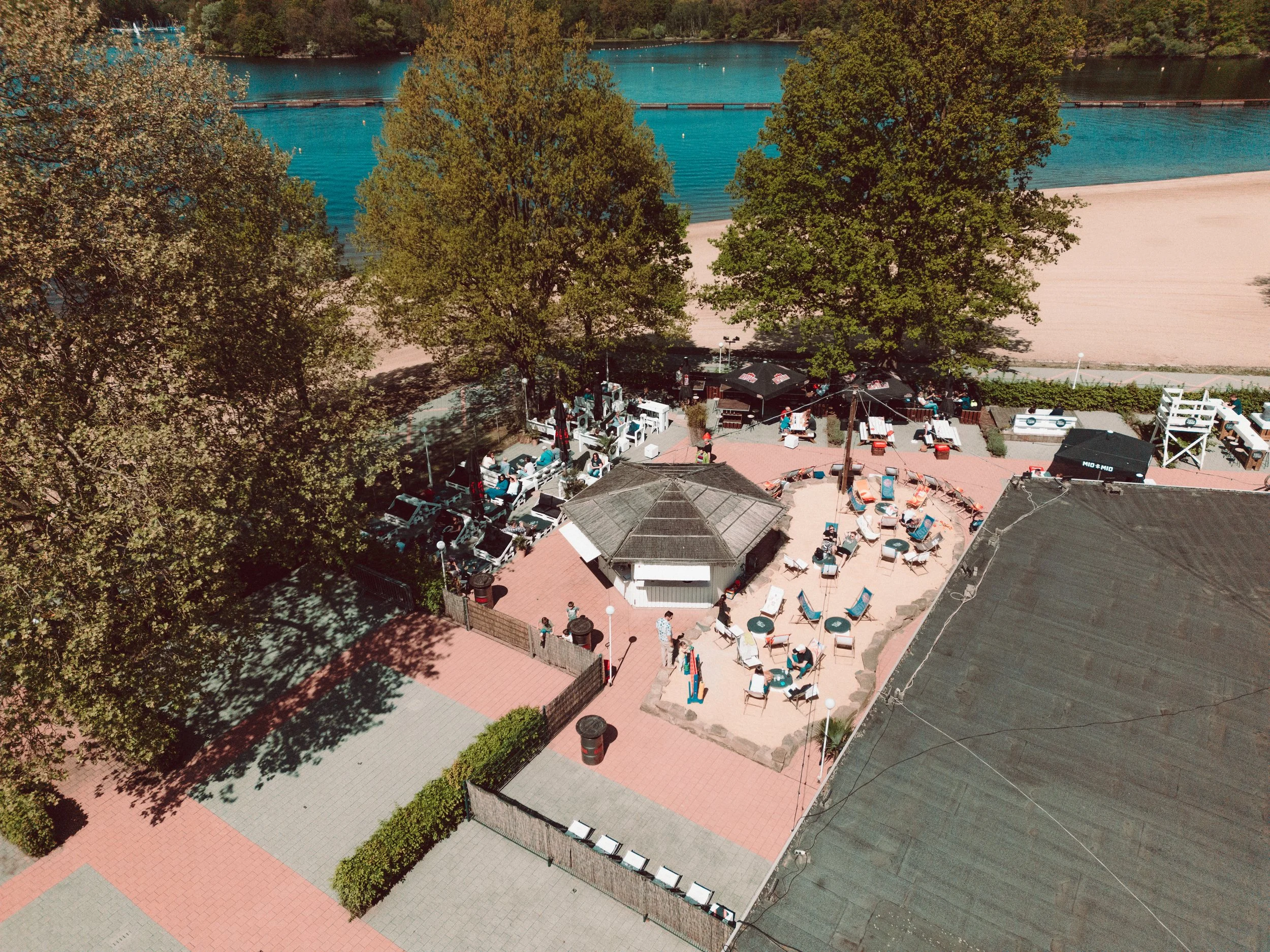 Luftaufnahme der Bar am Wasser mit Tischen, Sonnenliegen und Bäumen, bei schönem Wetter.