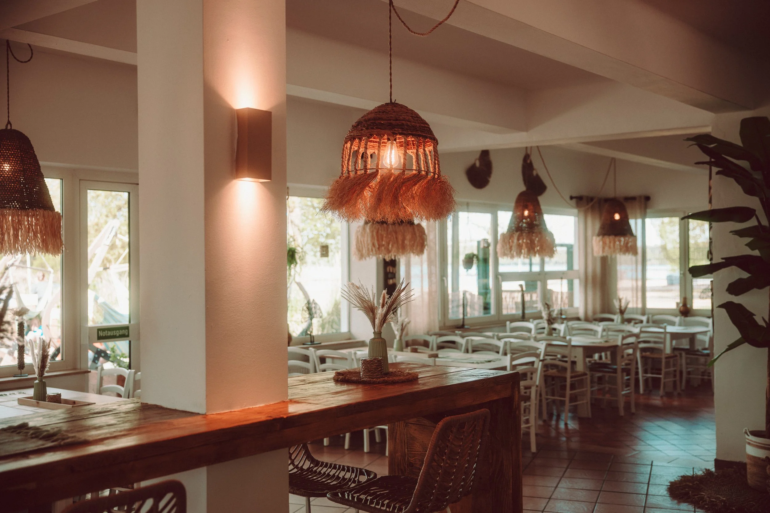 Ein gemütlich eingerichteter Restaurantraum mit weißen Stühlen, großen Fenstern, dekorativen Lampen und einer rustikalen Holztheke in warmem Licht.