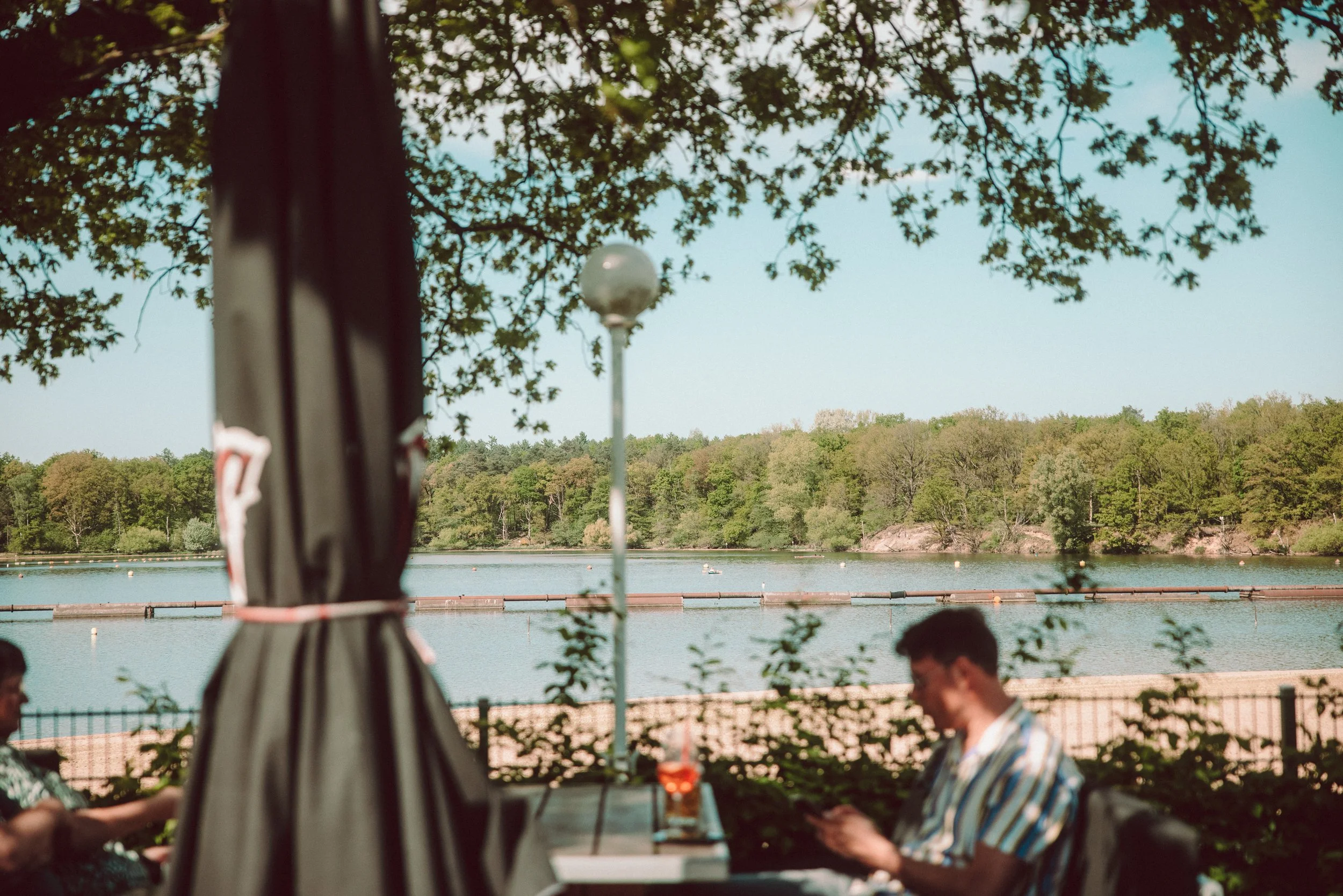 Menschen sitzen an einem Tisch im Freien unter Bäumen mit Blick auf den See, im Hintergrund sind Wasservögel und der Wald auf der gegenüberliegenden Seeseite zu erkennen