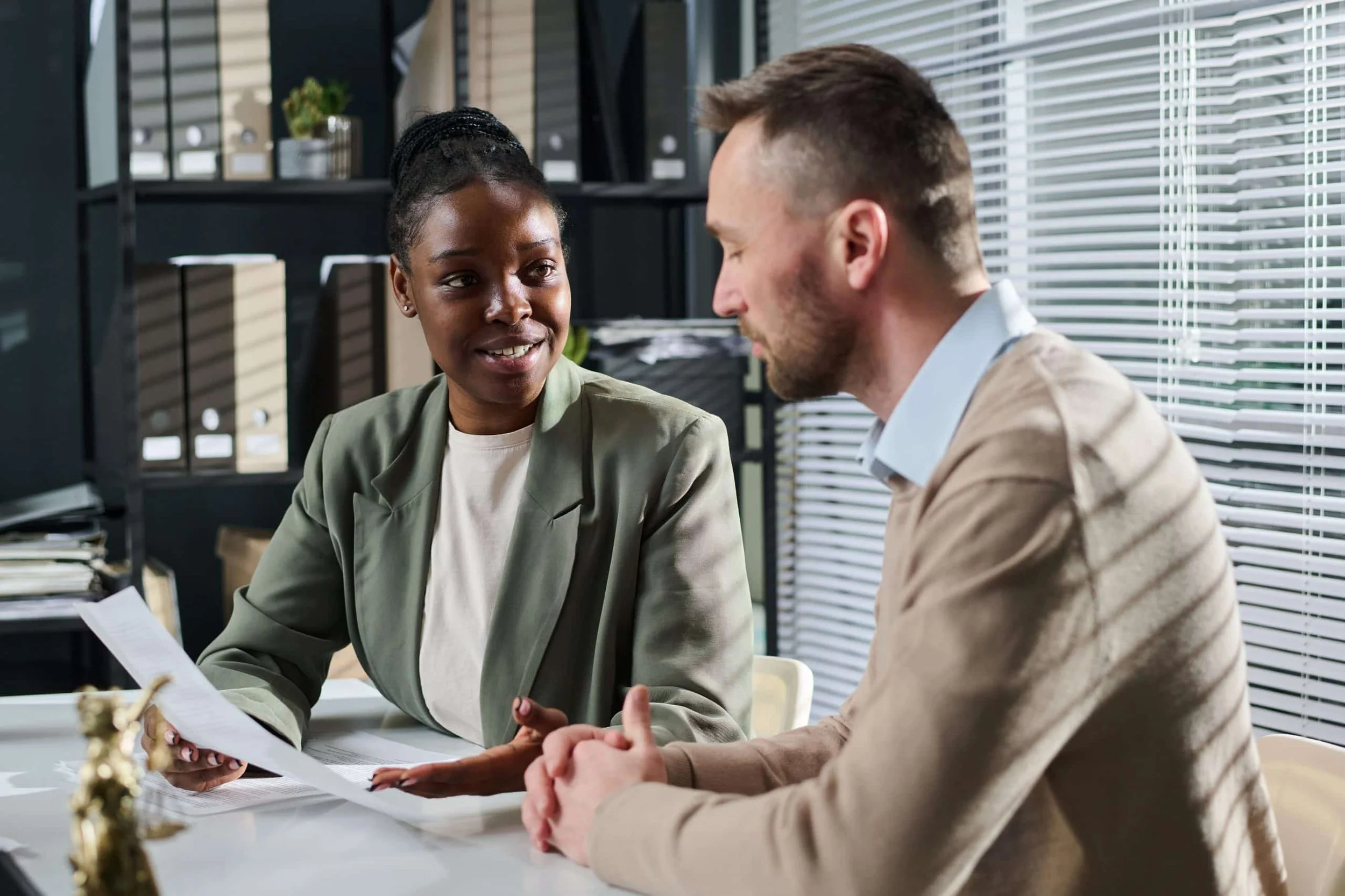 A woman and man having a conversation in an office, the woman is holding documents and smiling while the man listens attentively.
